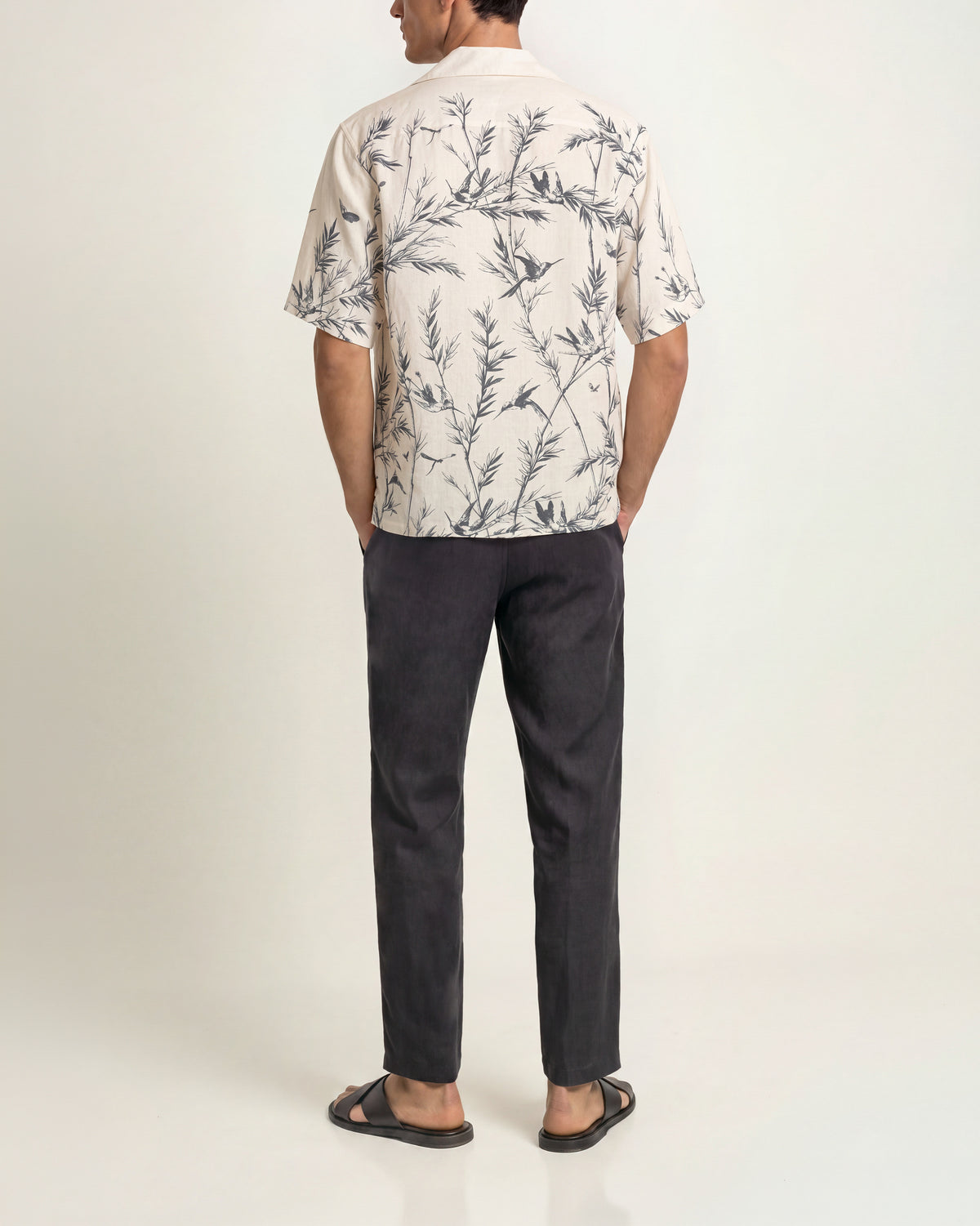 Back view of a man wearing a white shirt with navy pattern paired with charcoal trousers and black sandals on a plain background 