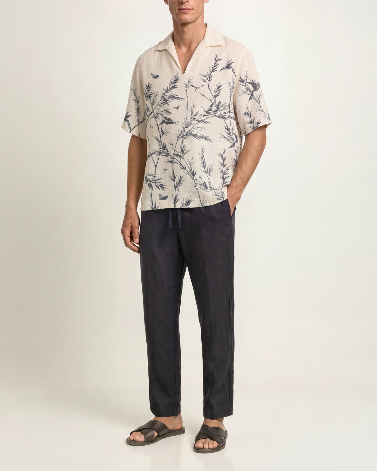 Man wearing a white with black pattern short-sleeved shirt paired with drawcord-waist dark navy trousers and brown sandals on a plain background