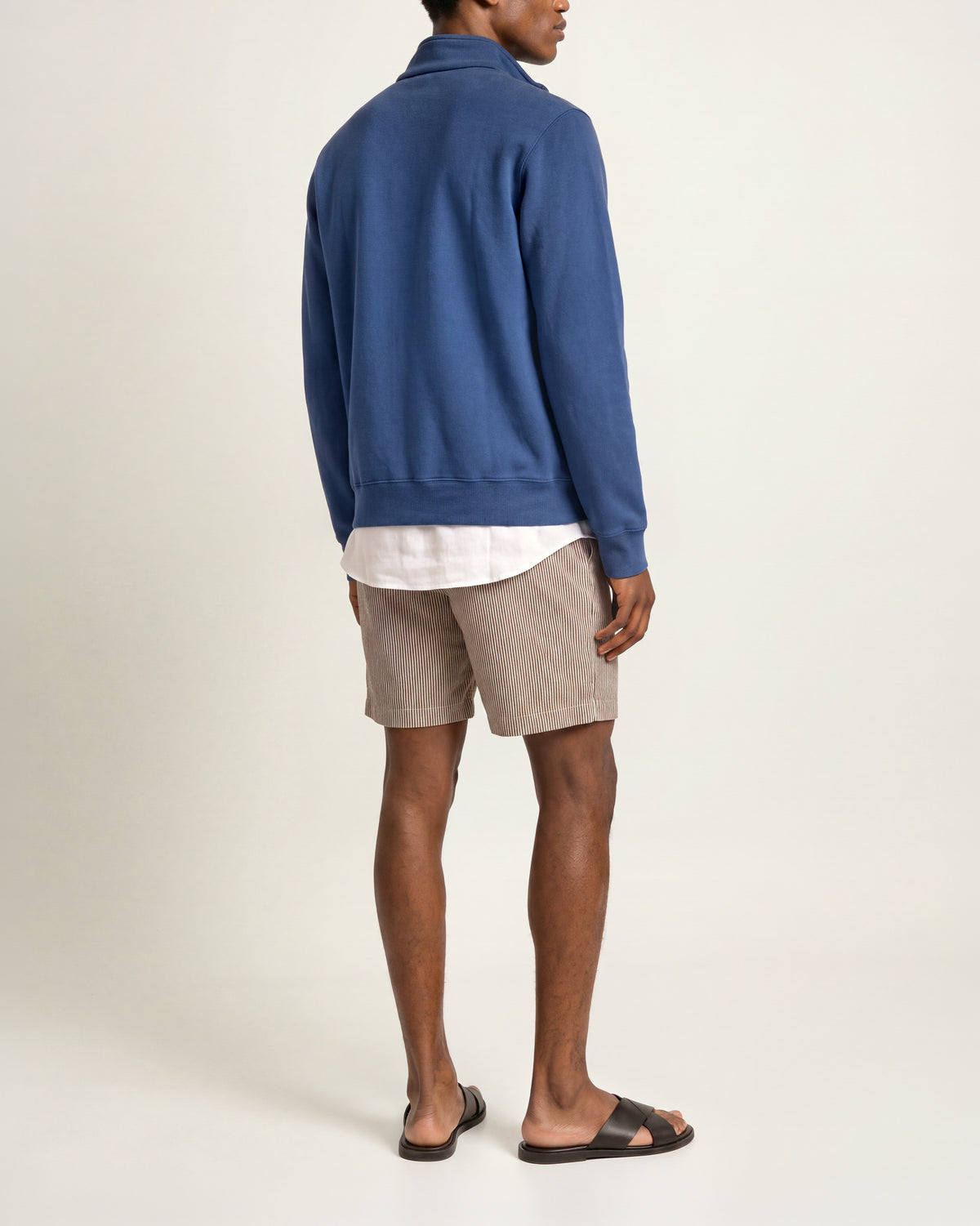 Man wearing dark blue sweatshirt with white t-shirt underneath and brown/ white striped shorts on a plain background
