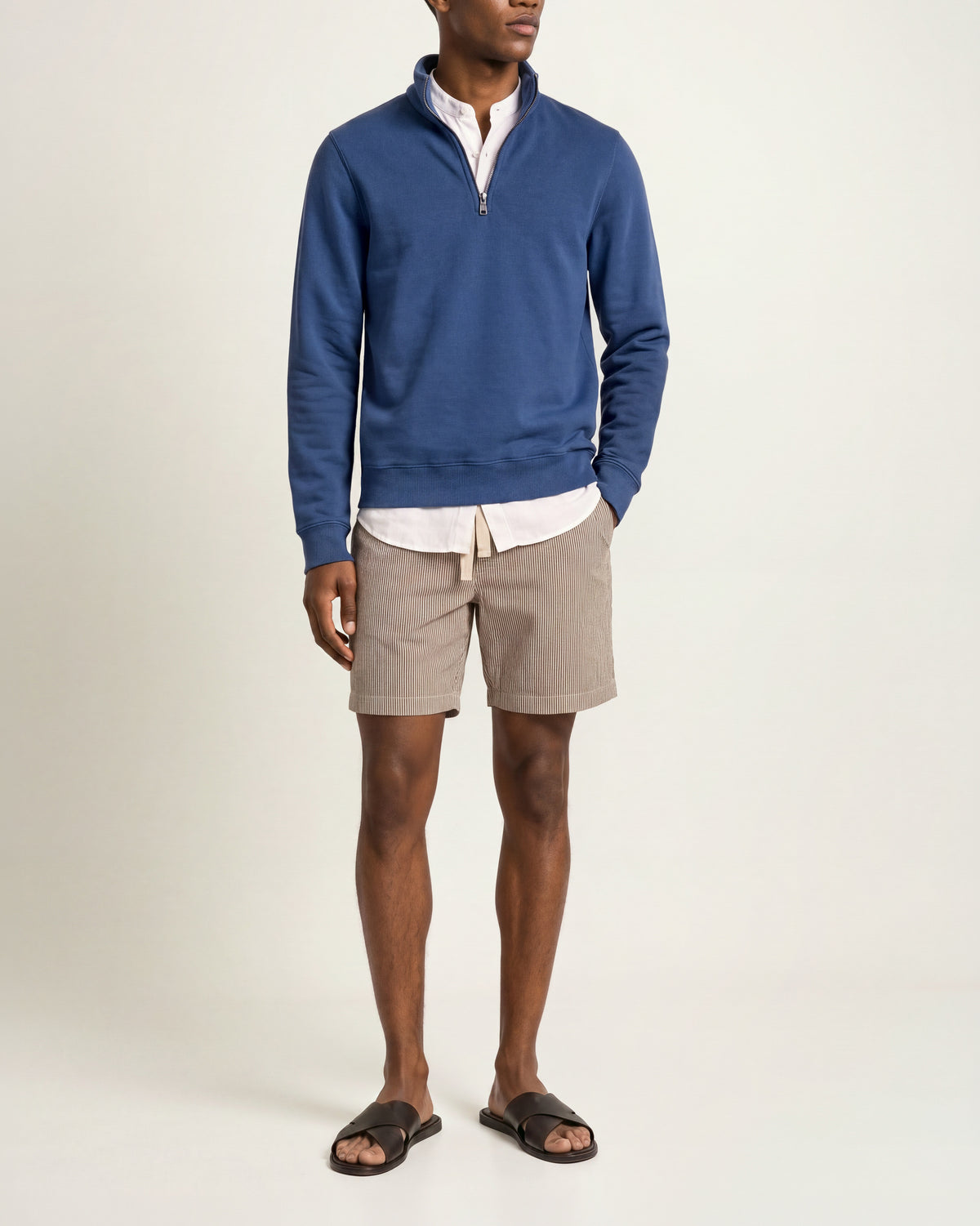 Man wearing dark blue sweatshirt with white t-shirt underneath and brown/ white striped shorts on a plain background