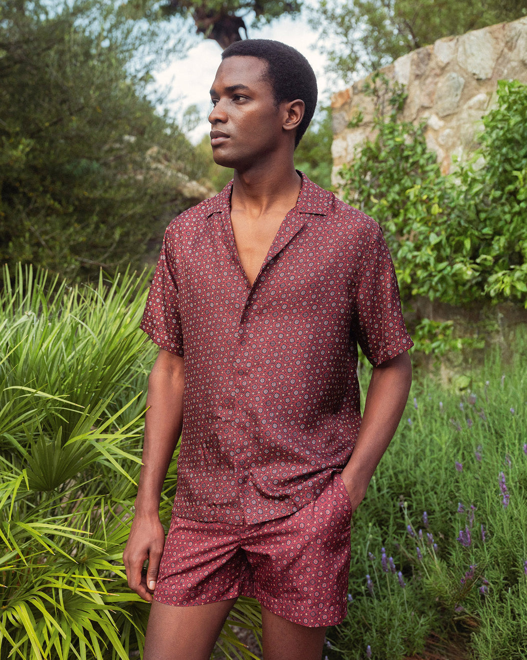 Man wearing a maroon patterned set standing in a garden.