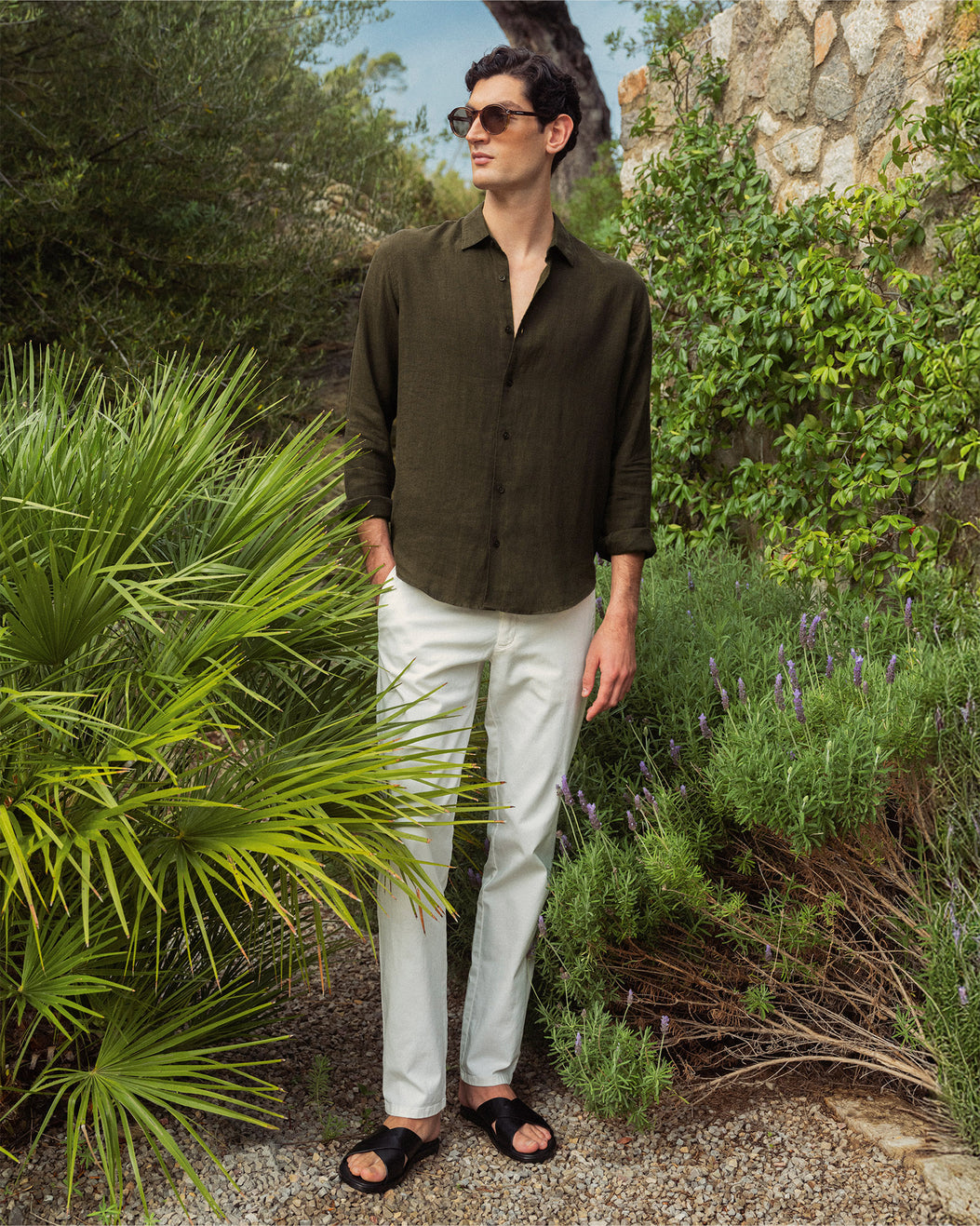 Man wearing khaki green shirt with white trousers standing amongst greenery