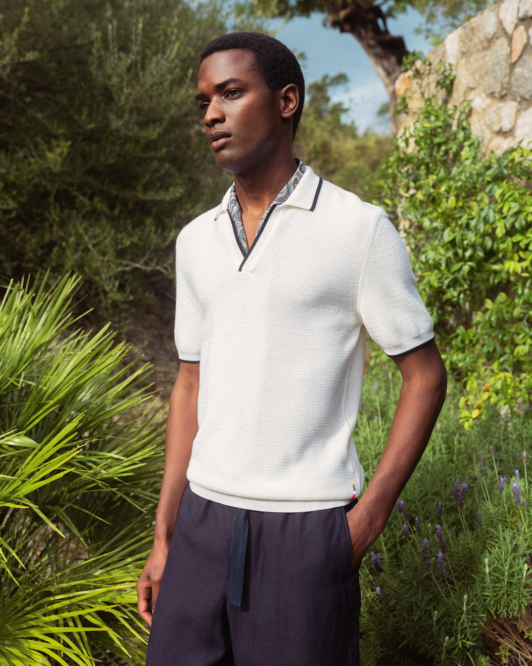 Man wearing a white polo shirt and dark pants standing in a natural setting with greenery.