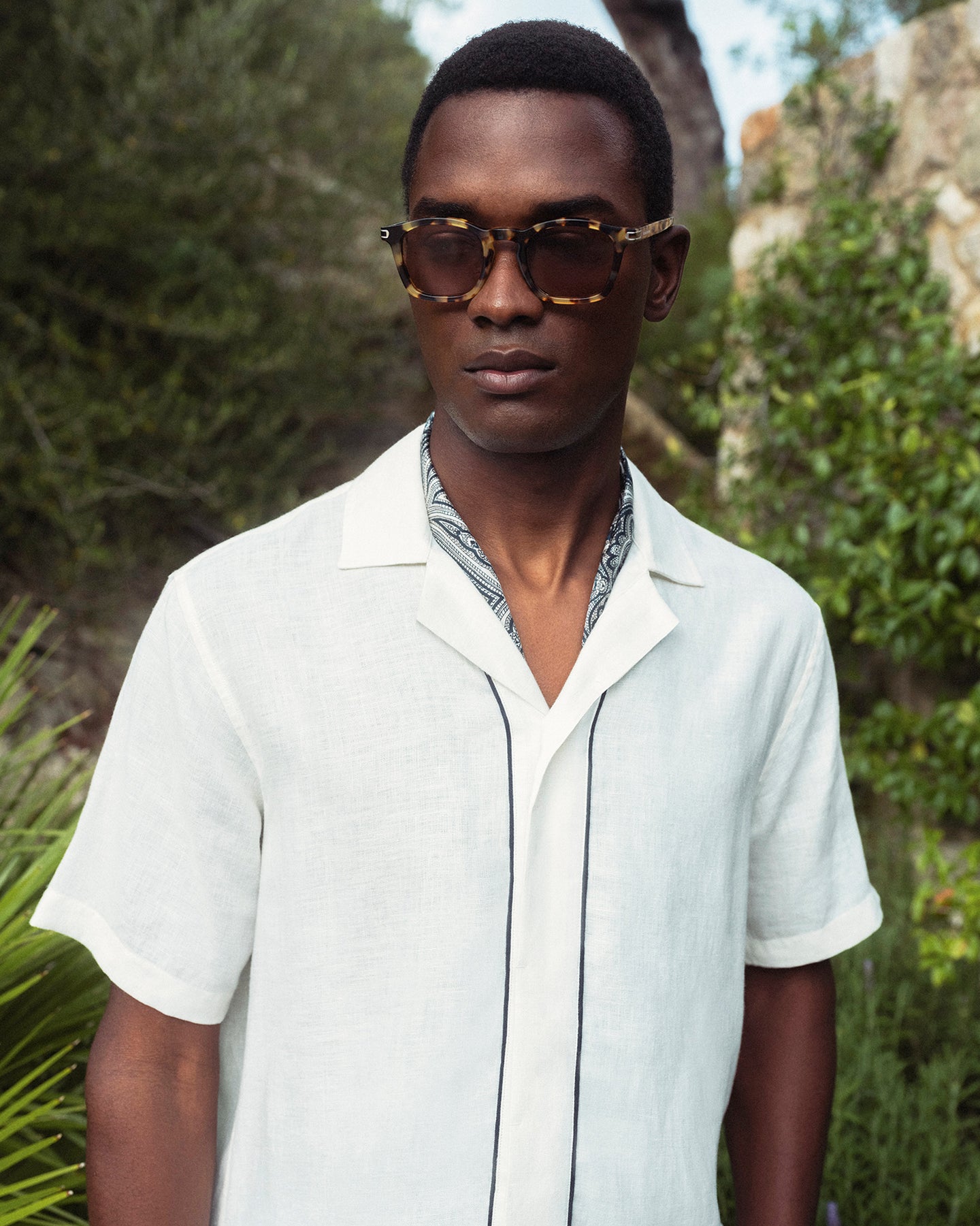 Man wearing sunglasses and a light-colored shirt outdoors with greenery in the background