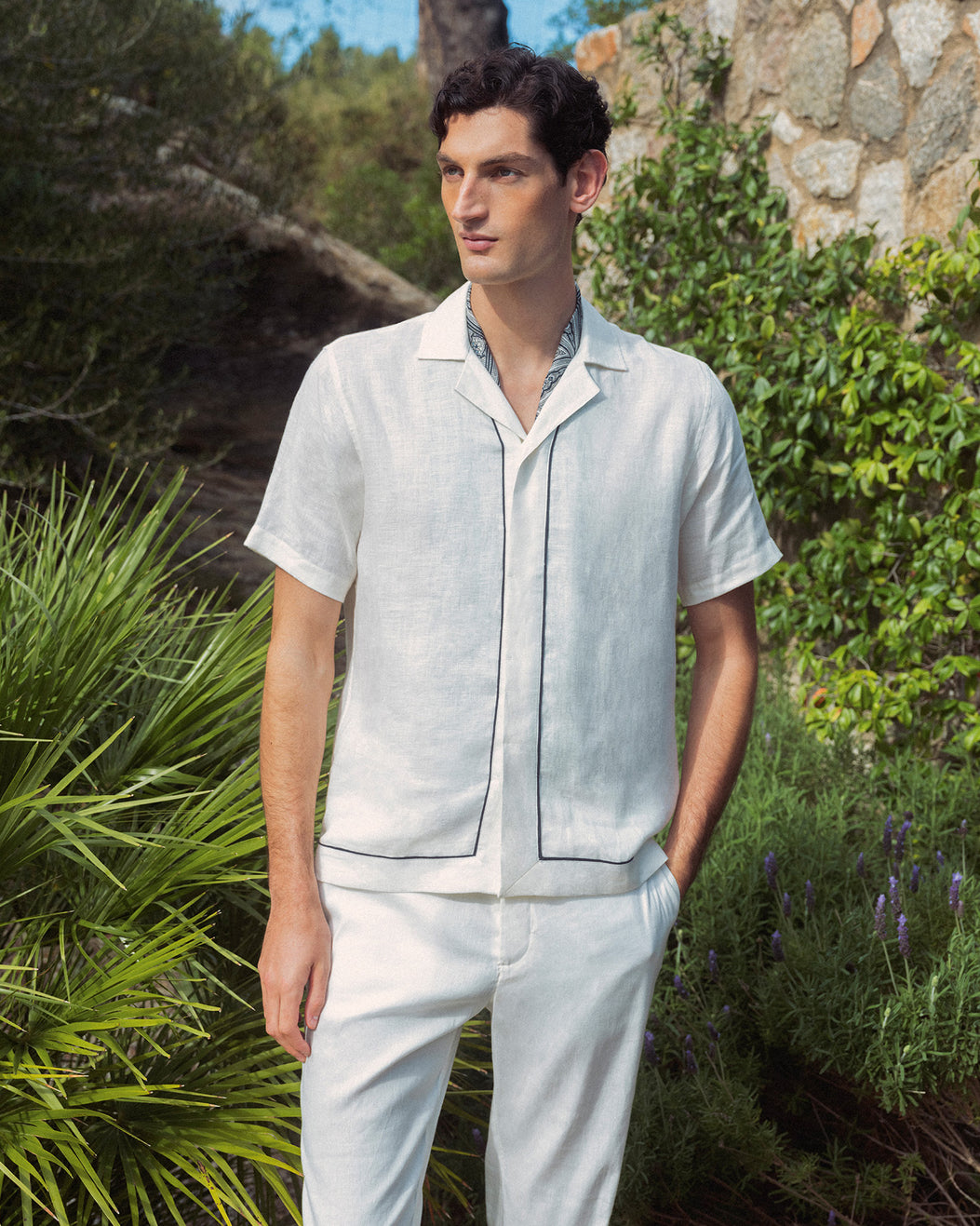 Man wearing white linen shirt with black piping with white trousers against greenery