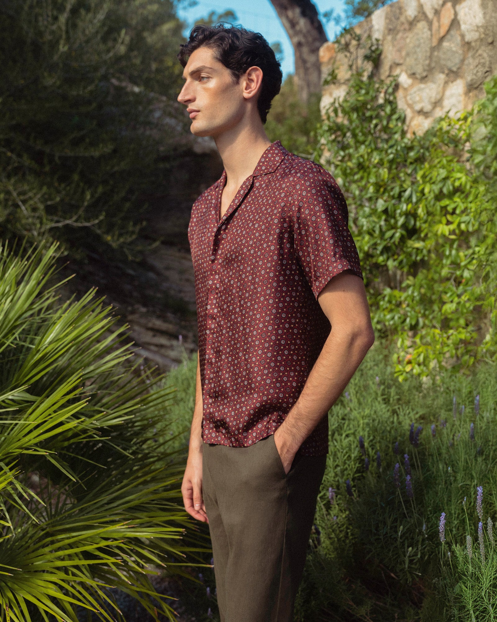 Man wearing a patterned shirt standing in a garden with greenery and a stone wall.