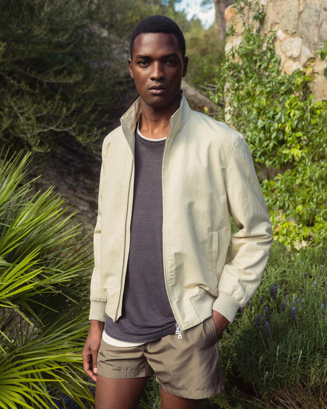 Man wearing a light jacket and shorts standing in a natural setting with greenery.