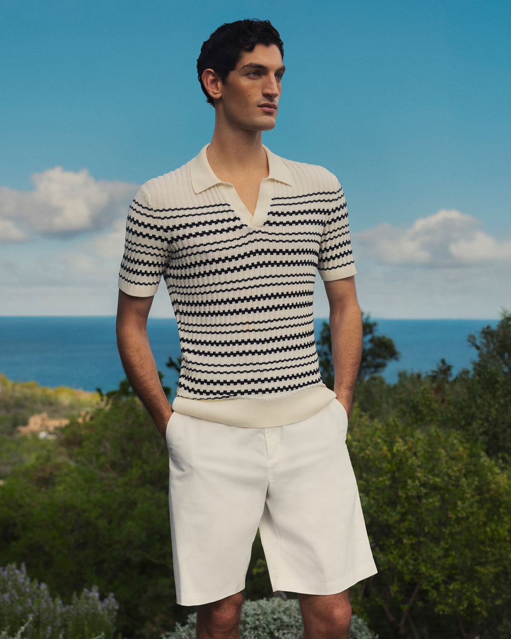 Man wearing a striped polo shirt and white shorts standing outdoors with a scenic background.