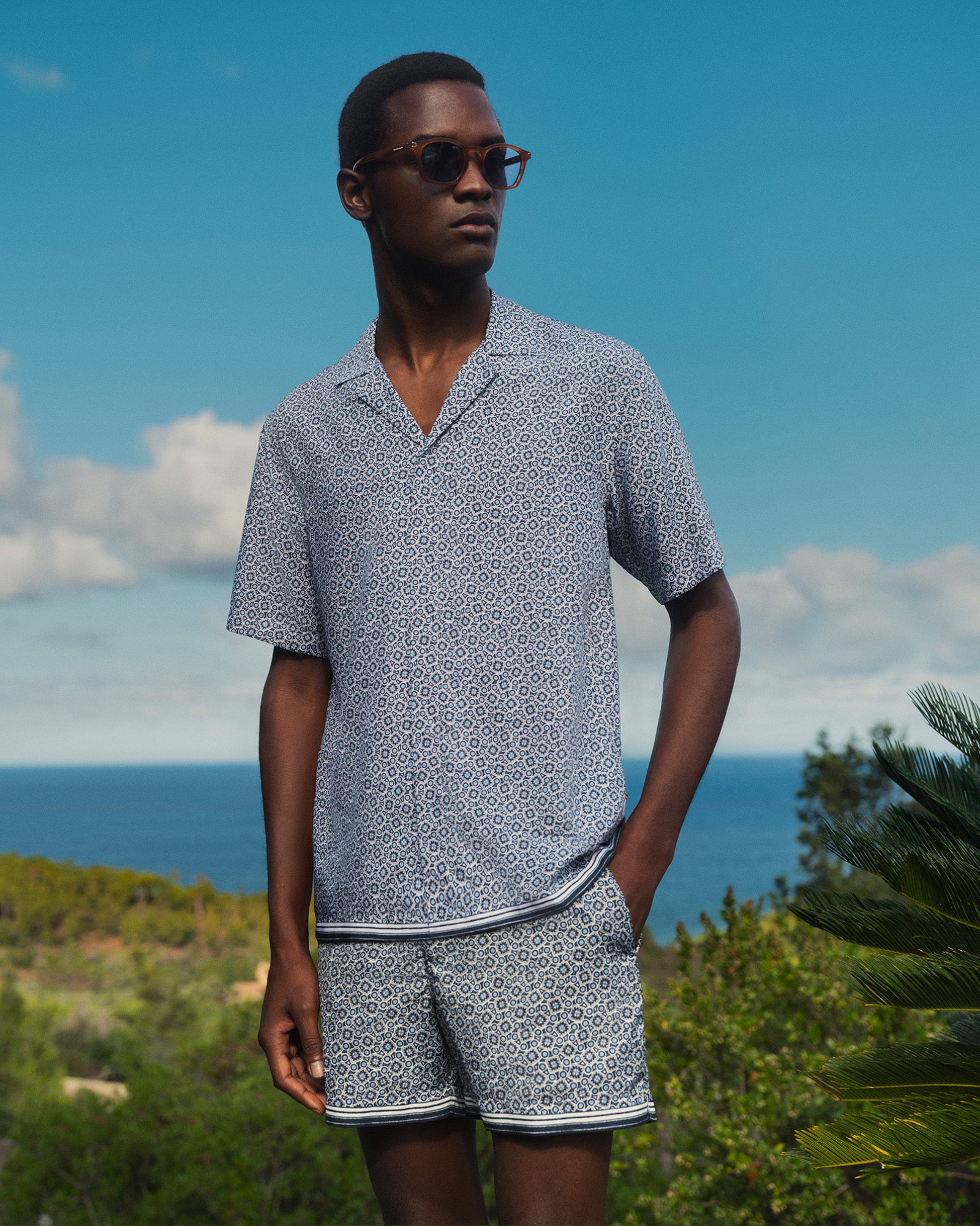 Man wearing a patterned shirt and shorts set with sunglasses, standing outdoors against a blue sky and ocean.