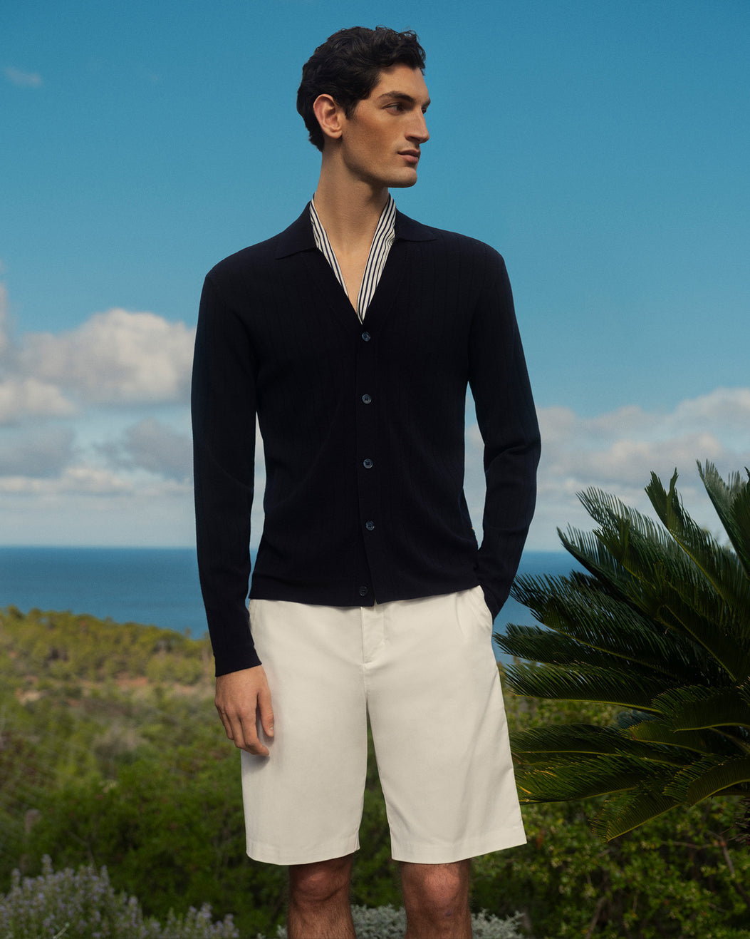 Man wearing a black cardigan and white shorts standing outdoors with a scenic background