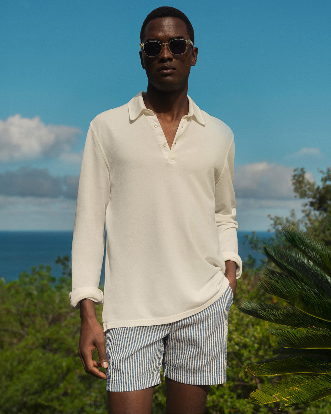 Man wearing a white shirt and striped shorts standing outdoors with a blue sky and ocean in the background