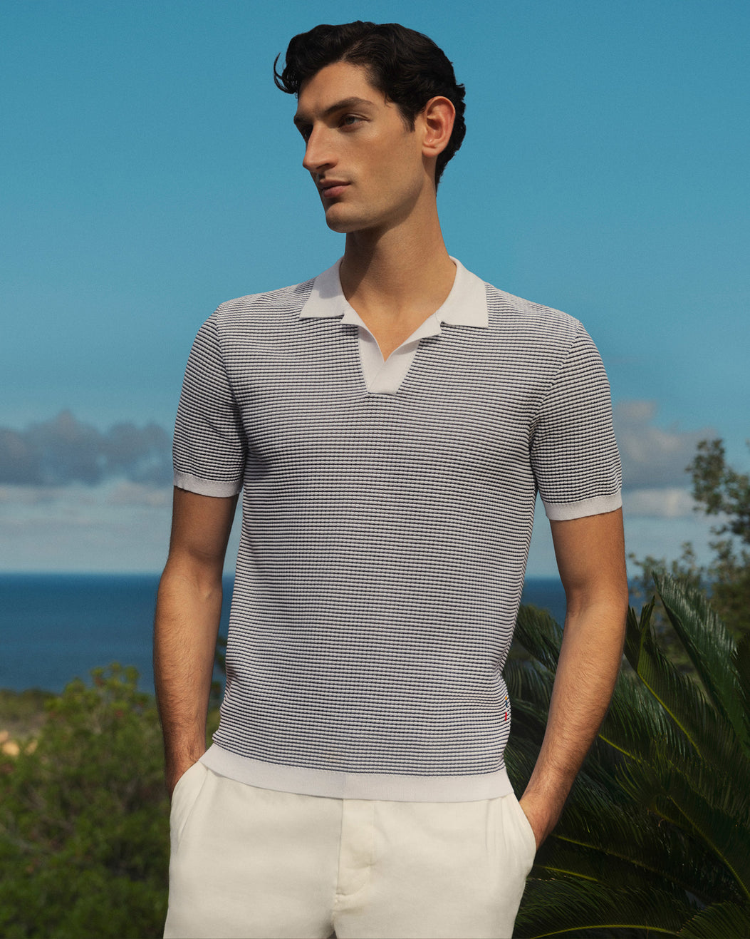Man wearing a striped polo shirt and white pants standing outdoors with a blue sky and ocean in the background