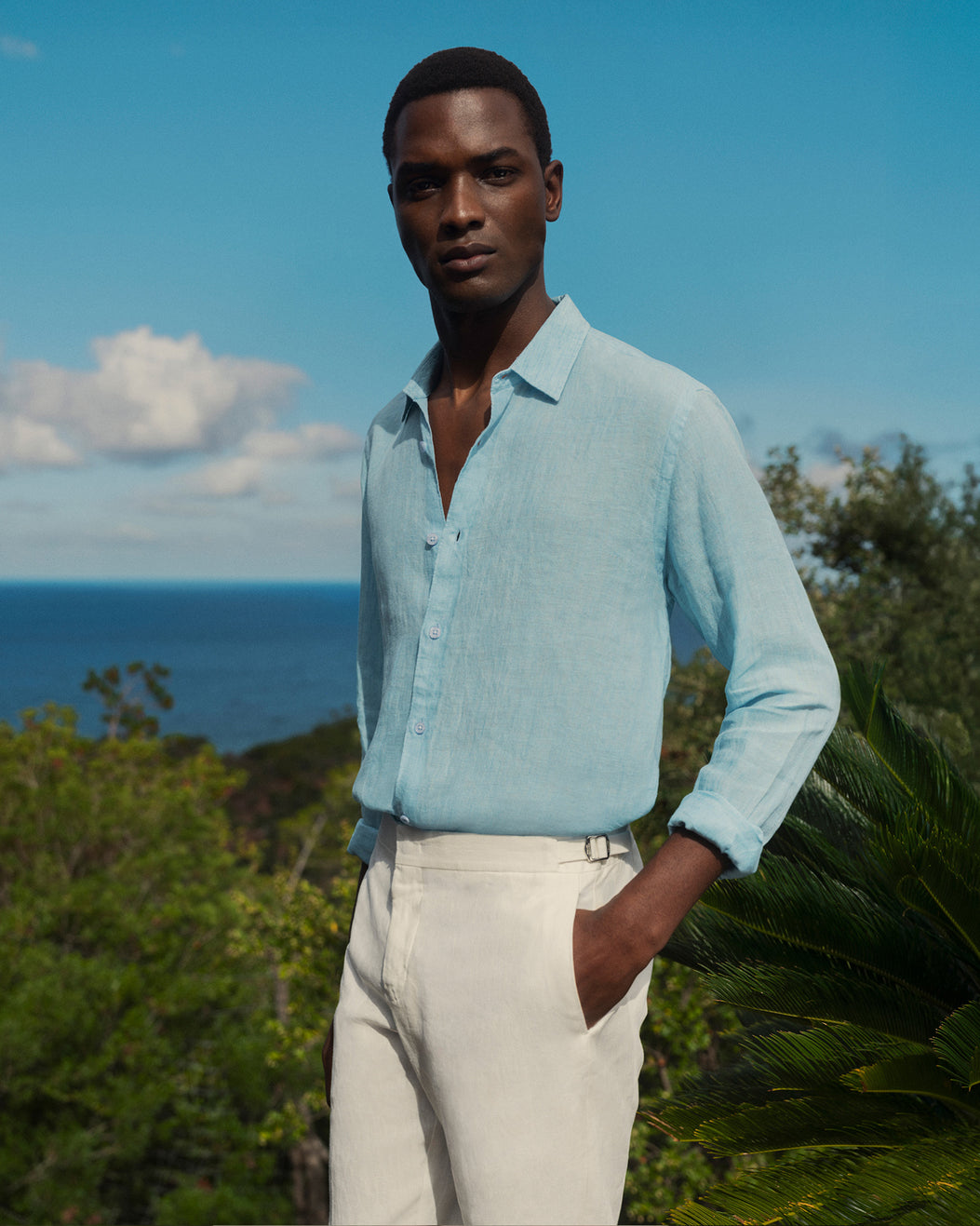Man wearing a light blue shirt and beige pants standing outdoors with ocean and sky in the background