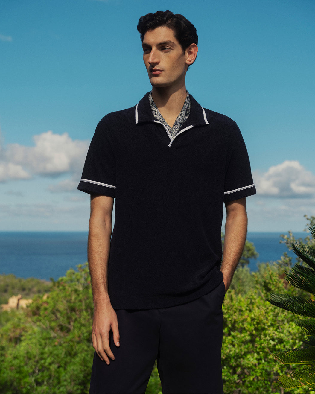 Man wearing a black polo shirt with white trim standing outdoors with ocean and sky in the background