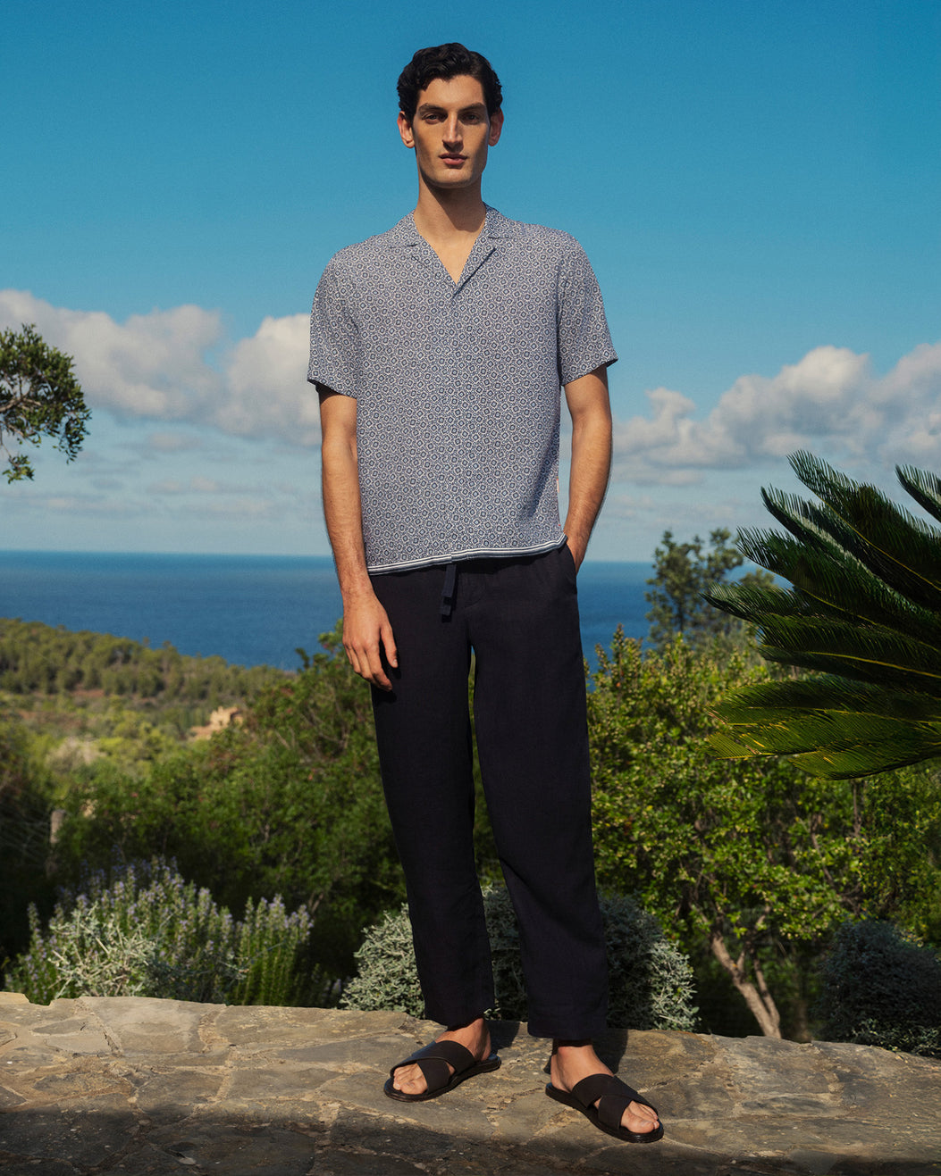 Man standing outdoors with ocean view wearing a patterned shirt and dark pants.