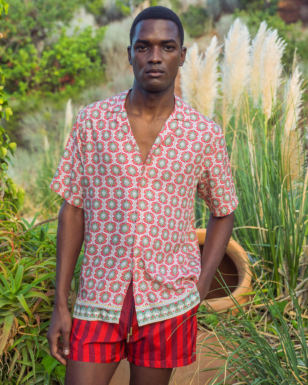 Man wearing a patterned shirt and red shorts standing in a natural setting with plants and a woven basket.