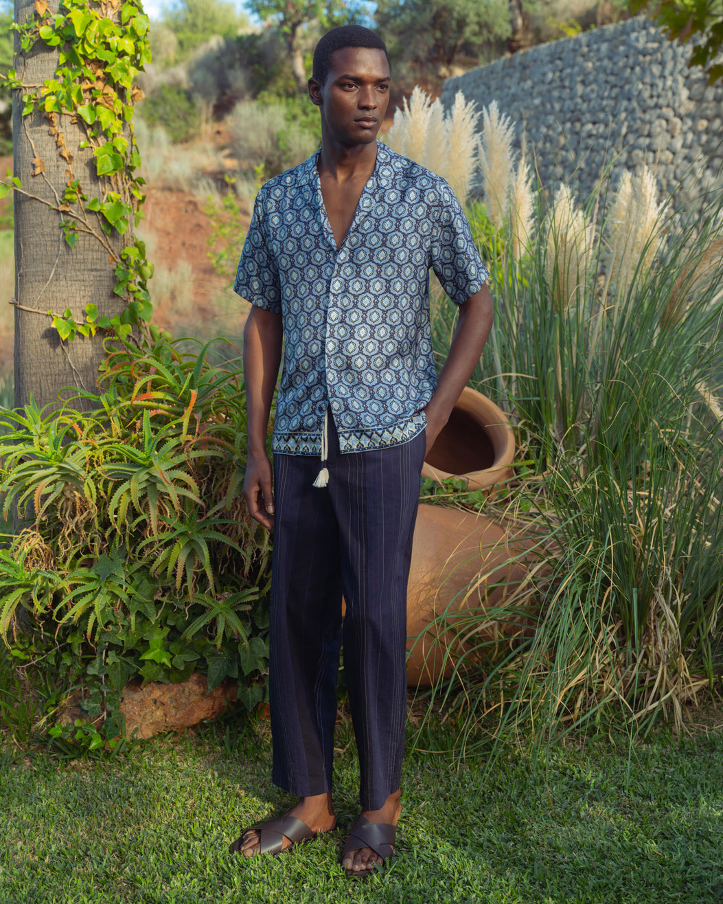Man wearing a blue patterned shirt and dark pants standing outdoors with greenery around