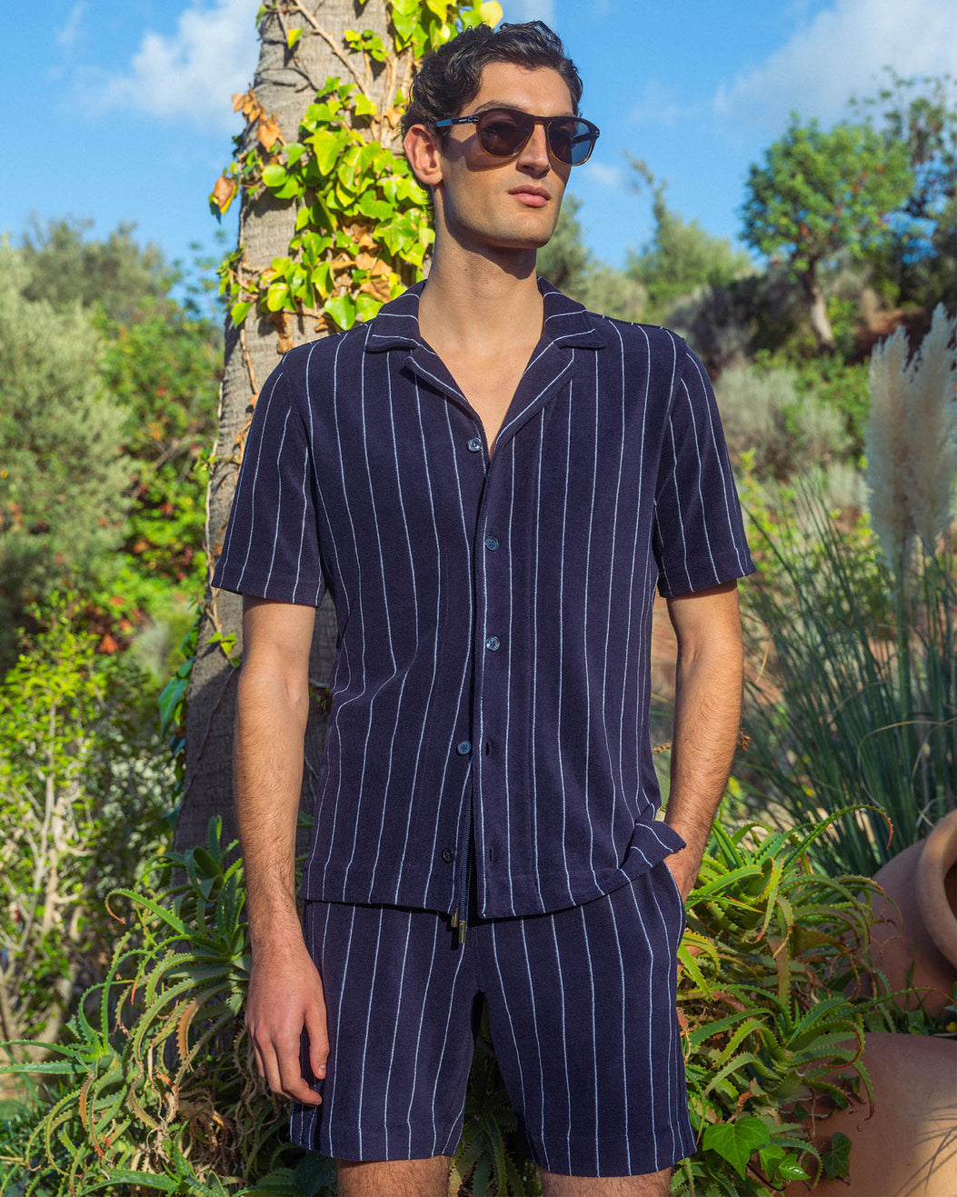 Man wearing a navy pinstripe outfit standing outdoors with greenery in the background