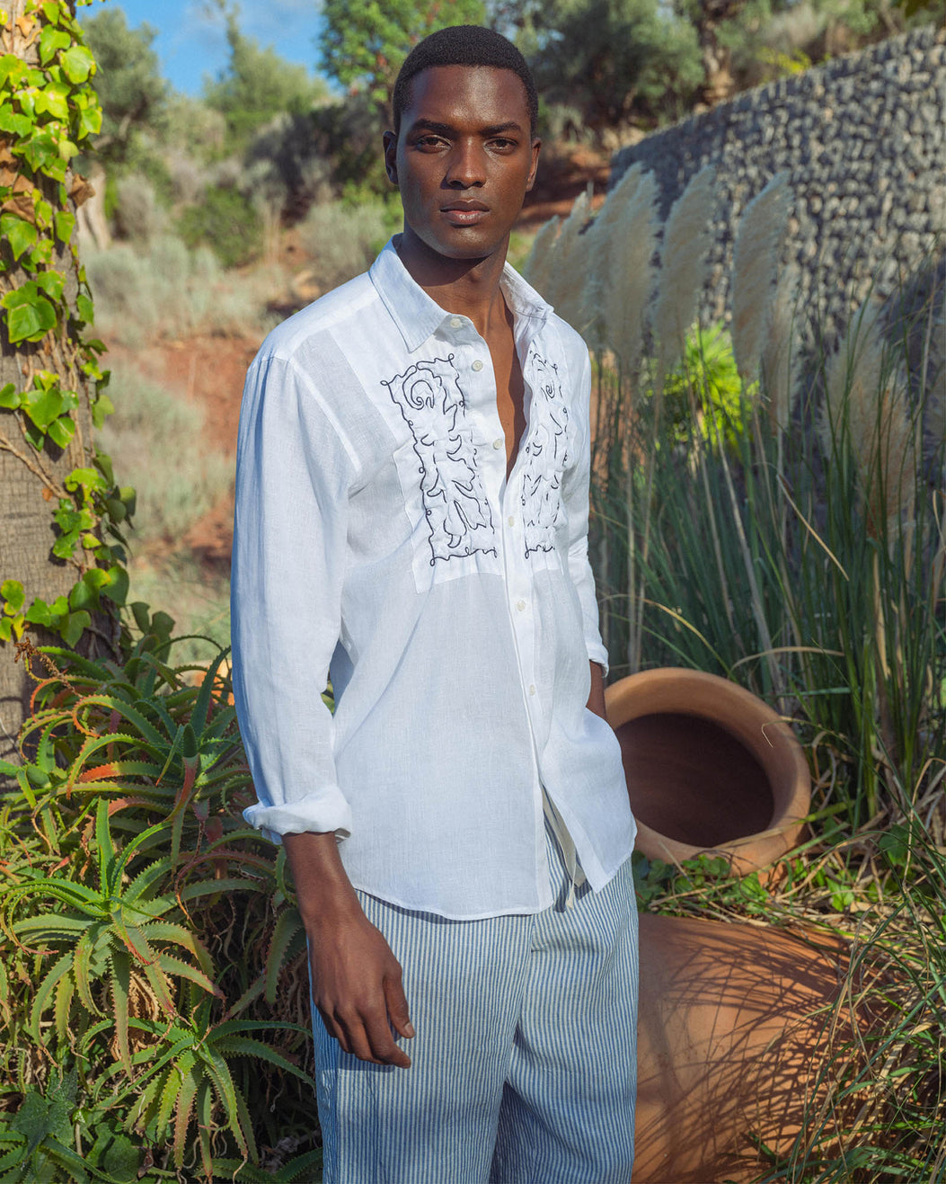 Man wearing a light blue embroidered shirt and striped pants standing outdoors with greenery.