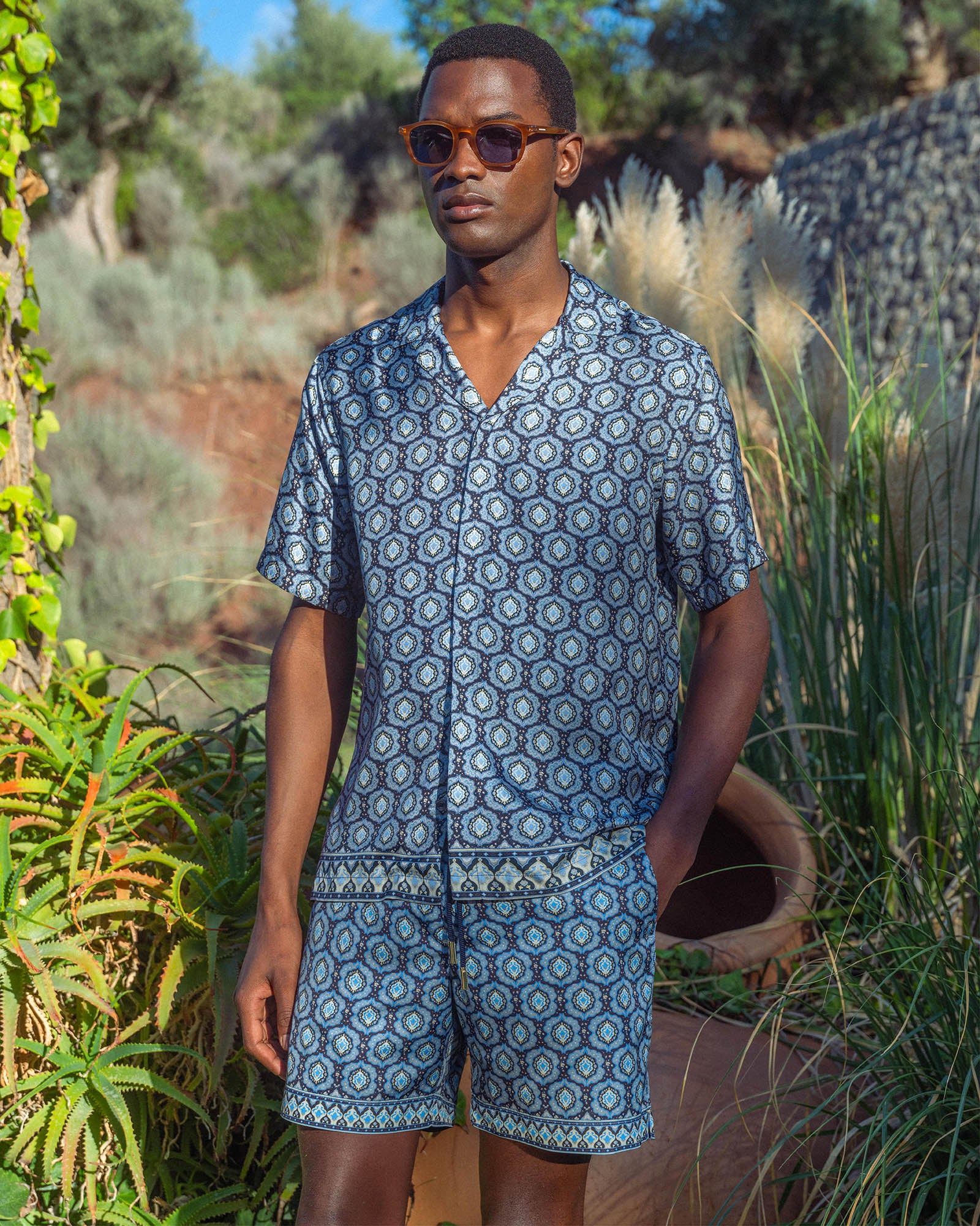 Man wearing a blue patterned shirt and shorts standing outdoors with greenery in the background
