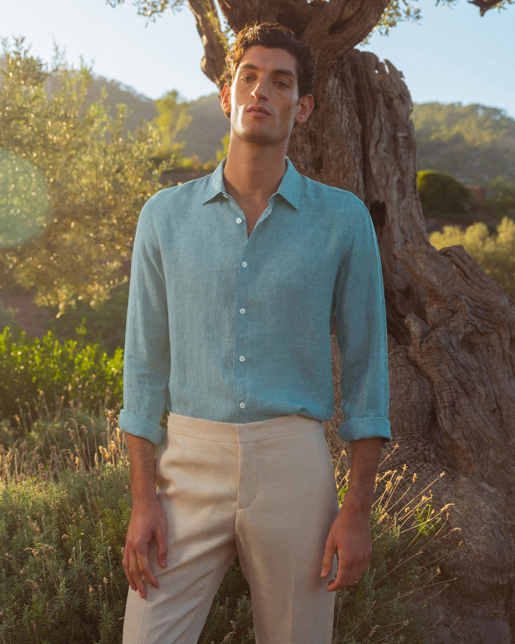 Man wearing light blue linen shirt with beige trousers against backdrop of trees
