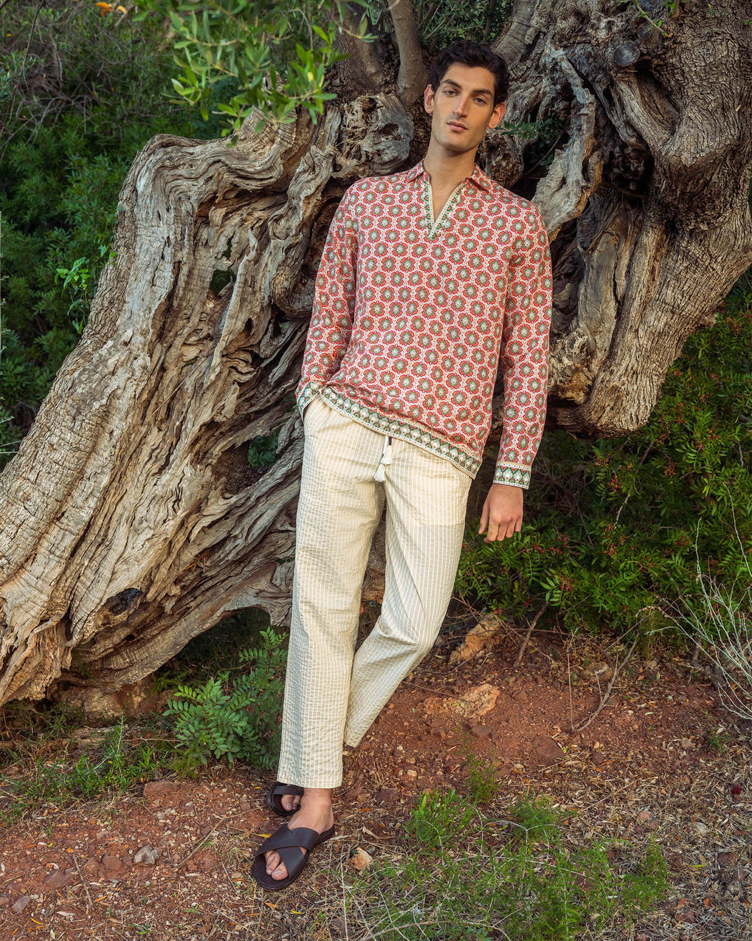 Man wearing a patterned shirt and light pants standing next to a large tree in a natural setting