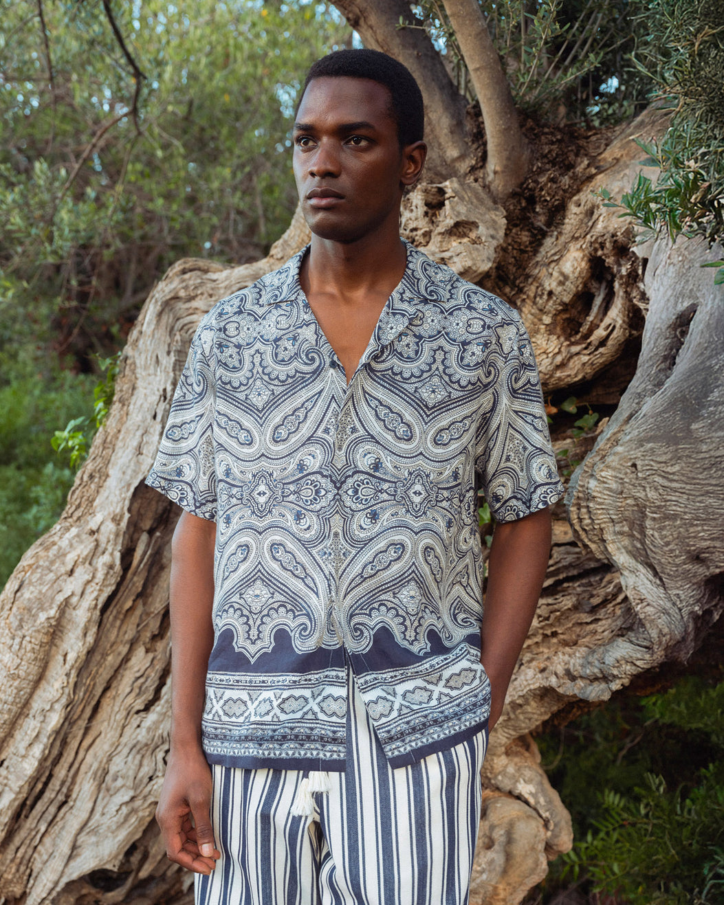 Man wearing a patterned shirt and striped pants standing in front of a large tree.