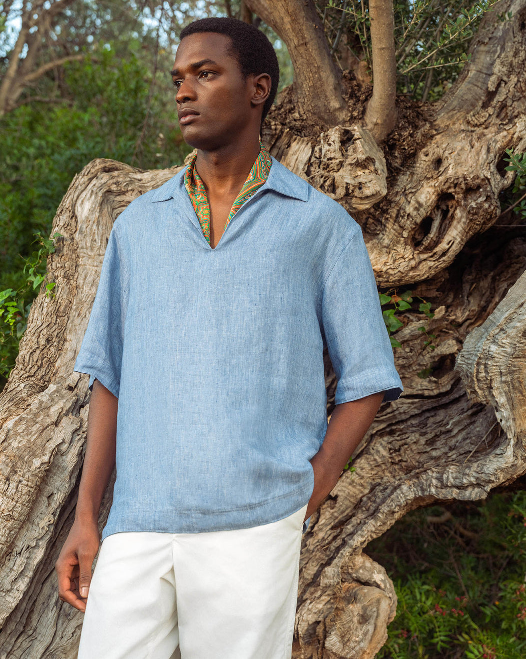 Man wearing a blue shirt and white pants standing against a large tree trunk.