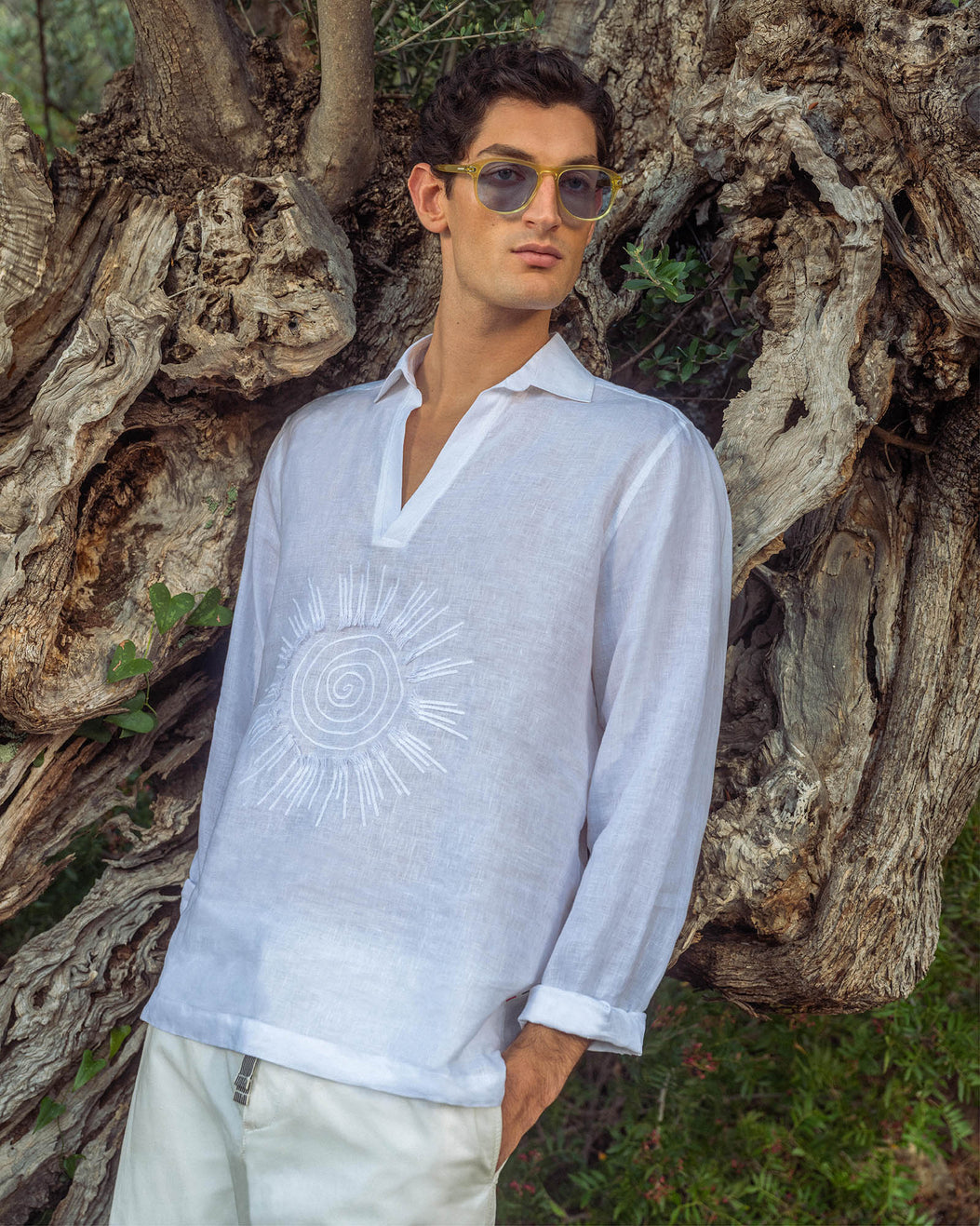 Man wearing white shirt with sunshine embroidery detailing, leaning against a tree
