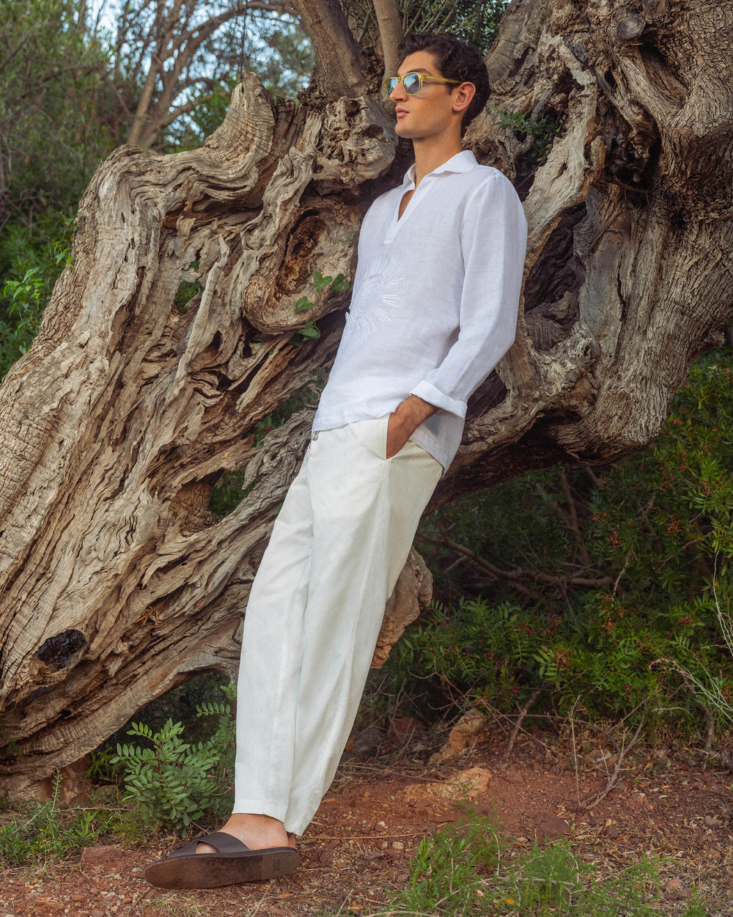 Man in white shirt and pants standing next to a large tree in a natural setting
