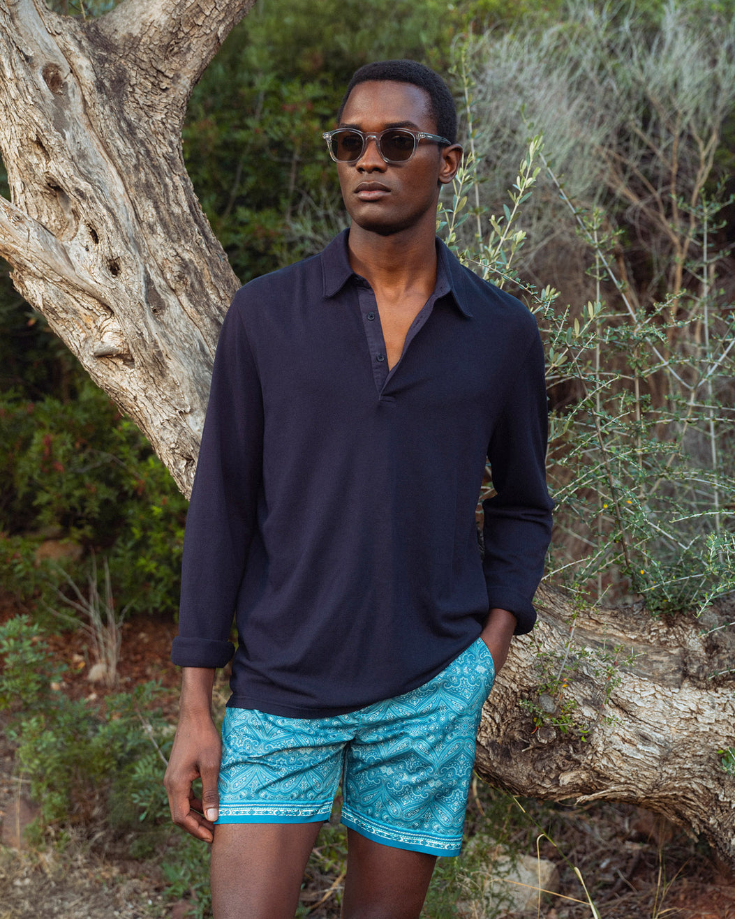 Man wearing a navy long-sleeve shirt and blue patterned shorts standing in a natural setting.