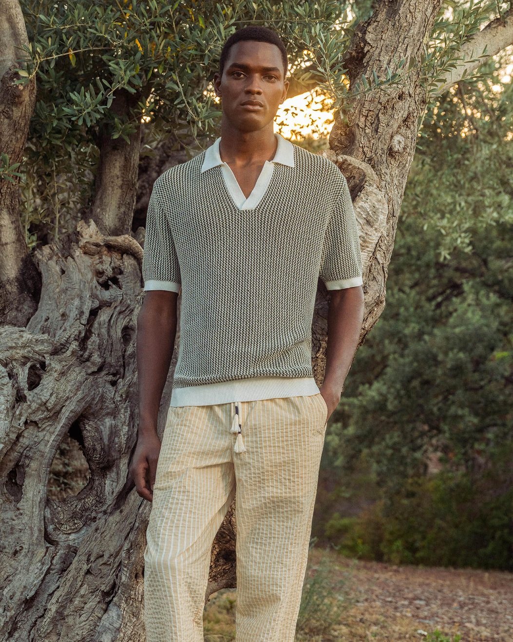 Man wearing a gray knit sweater and beige pants standing in a natural setting with trees.