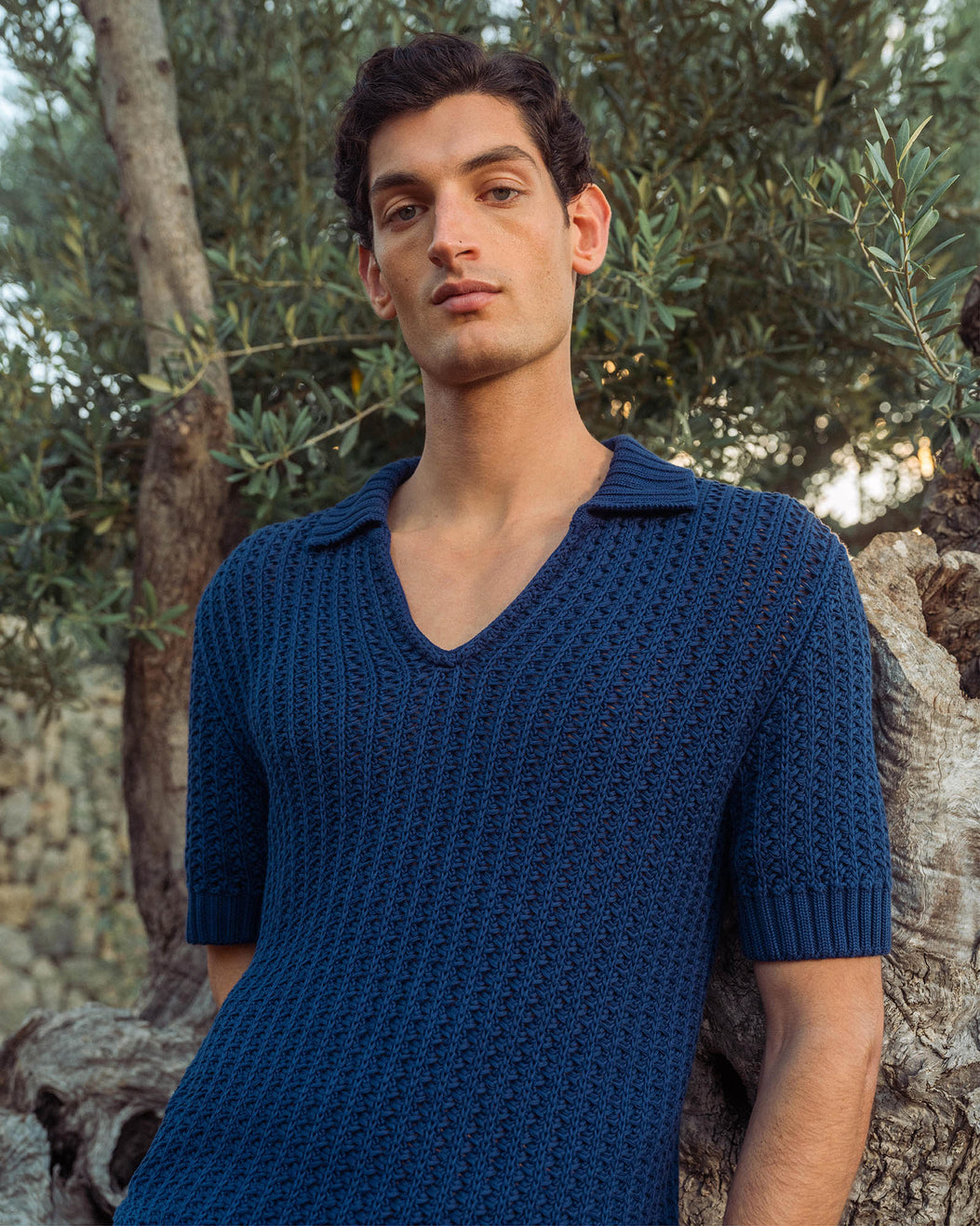Man wearing a blue knitted shirt standing outdoors with greenery in the background