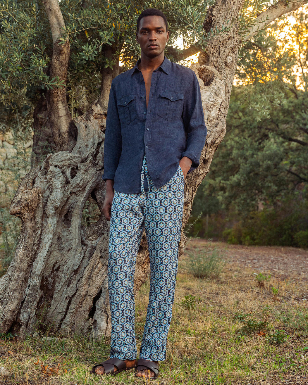 Man wearing a blue shirt and patterned pants standing in a natural setting with trees.