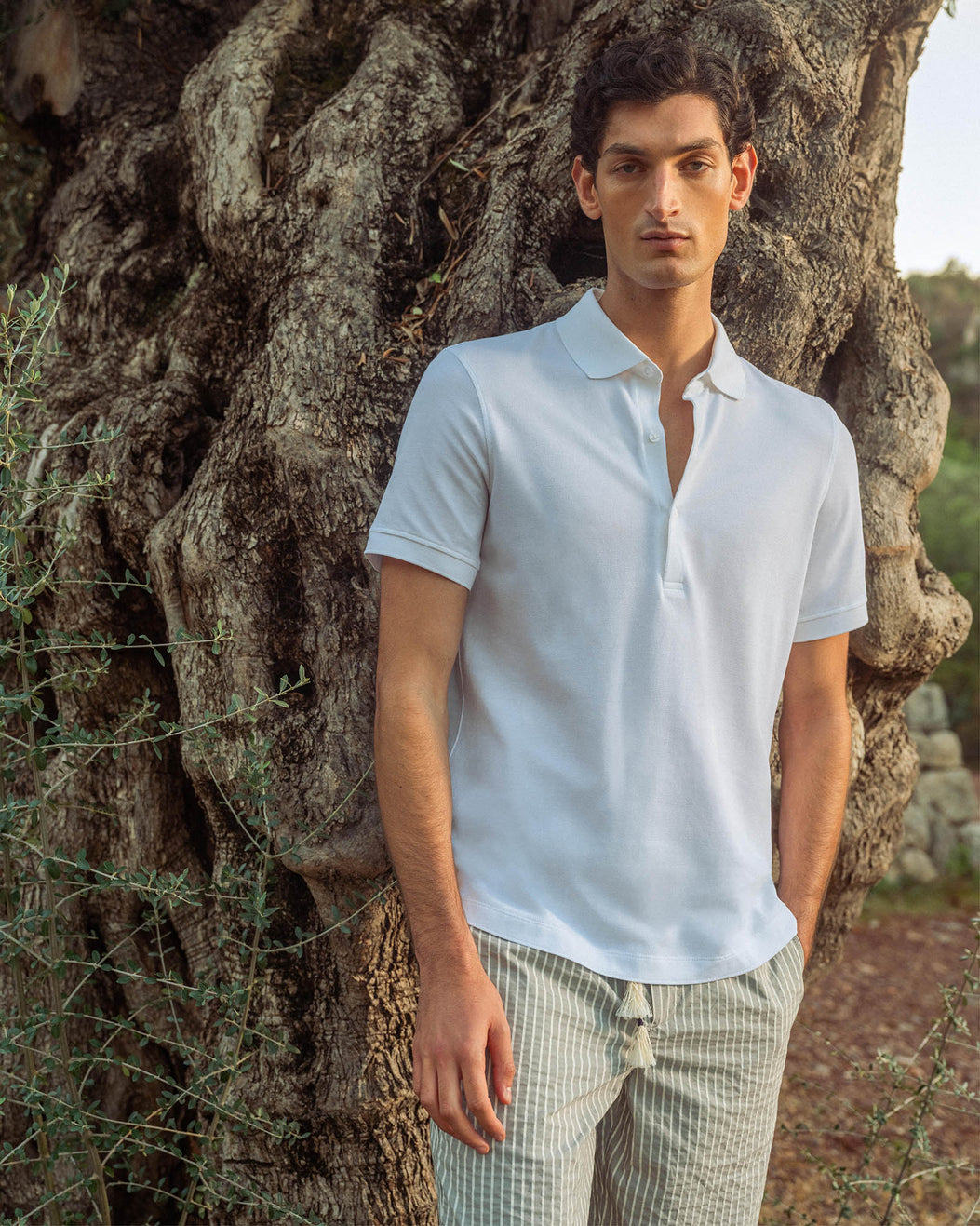 Man wearing a white polo shirt and plaid pants standing against a textured tree.