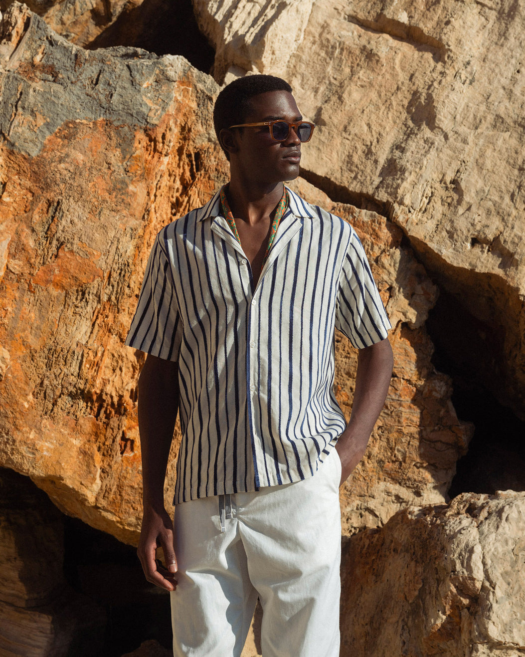 Man wearing a striped shirt and sunglasses standing against a rocky background