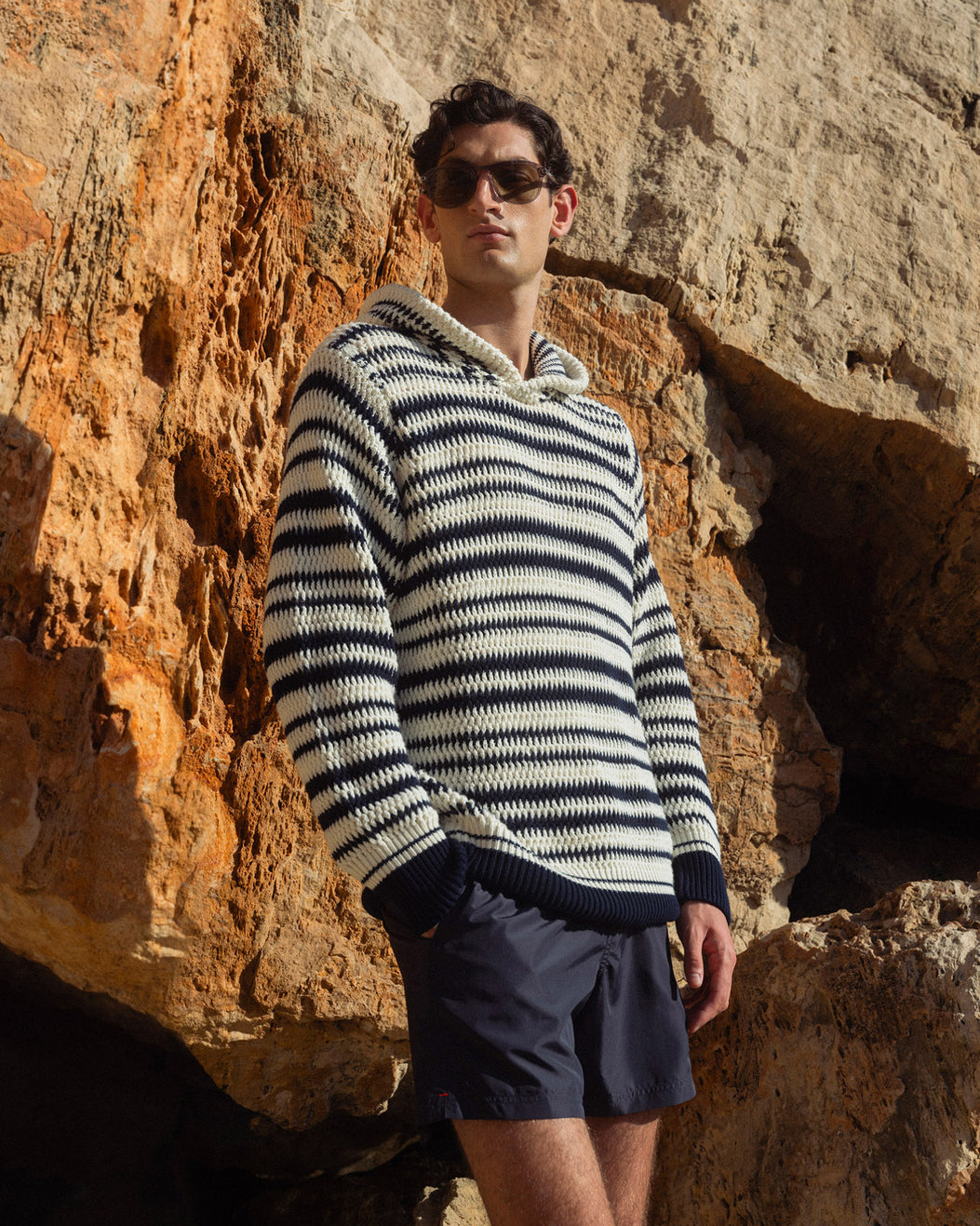 Man wearing a striped sweater and sunglasses standing against a rocky wall.
