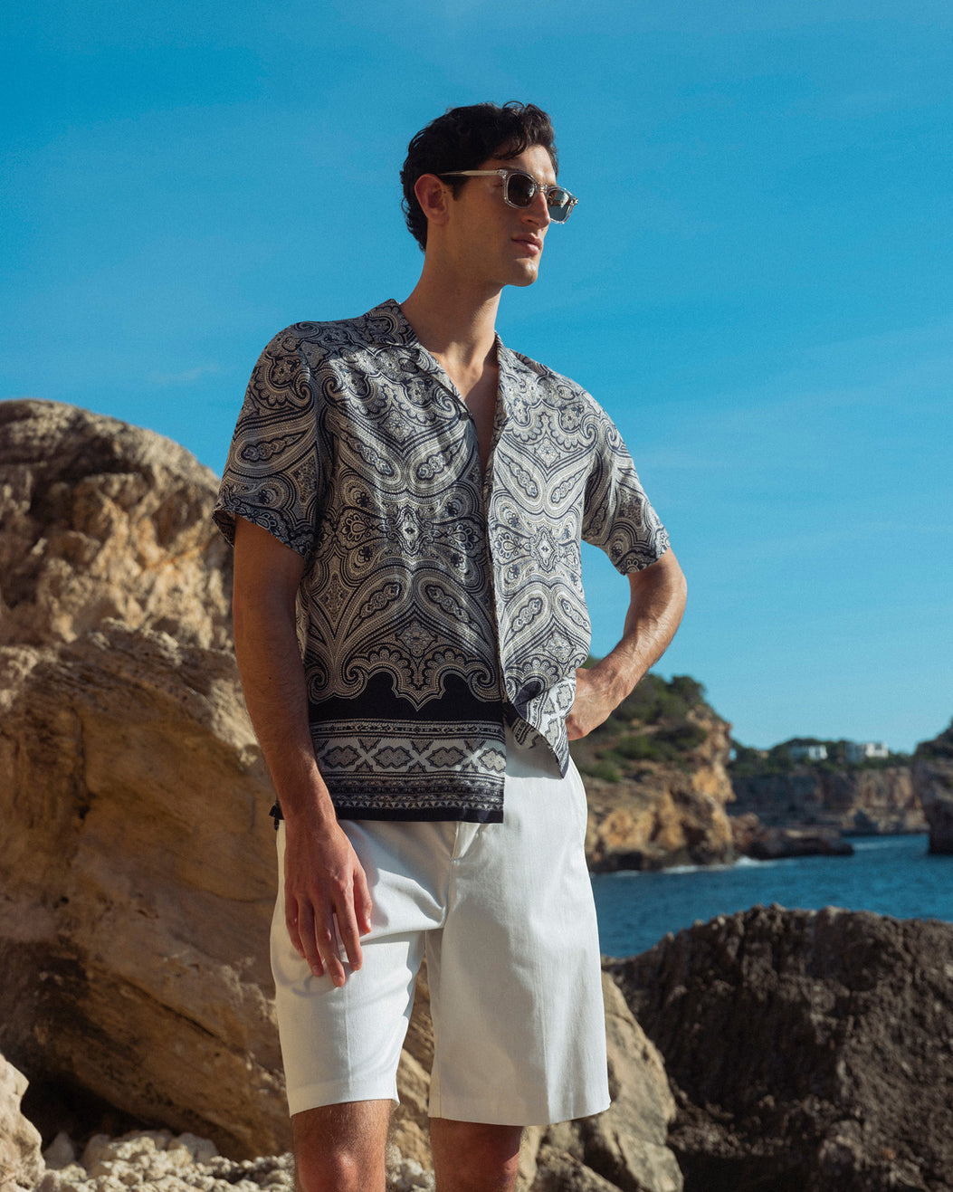 Man wearing a patterned shirt and sunglasses standing on rocky terrain with ocean and cliffs in the background.