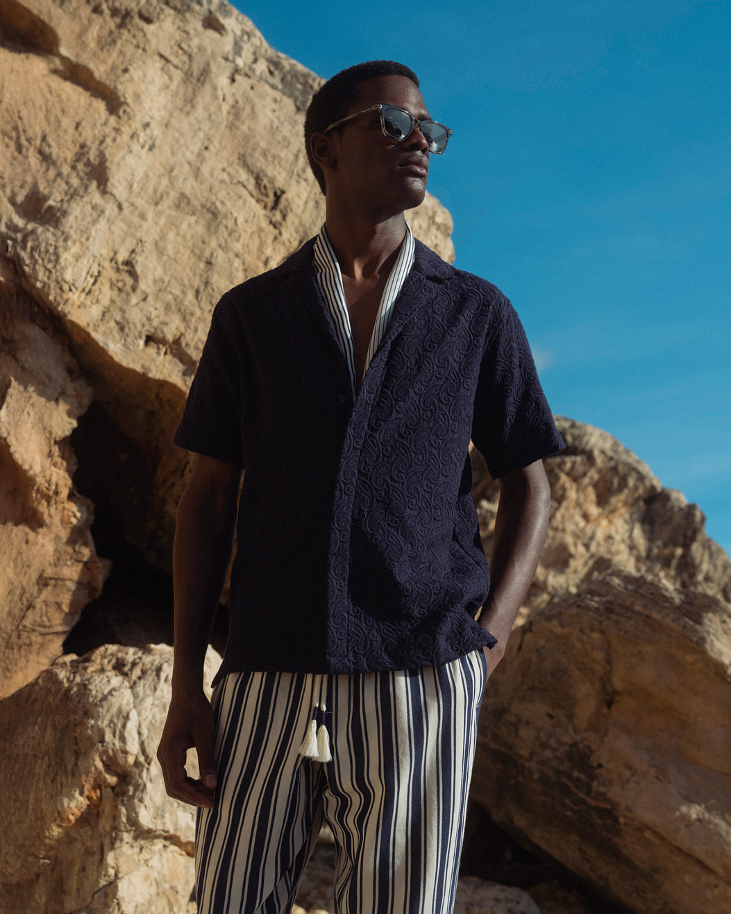 Man wearing sunglasses and a dark shirt with striped pants standing against a rocky background