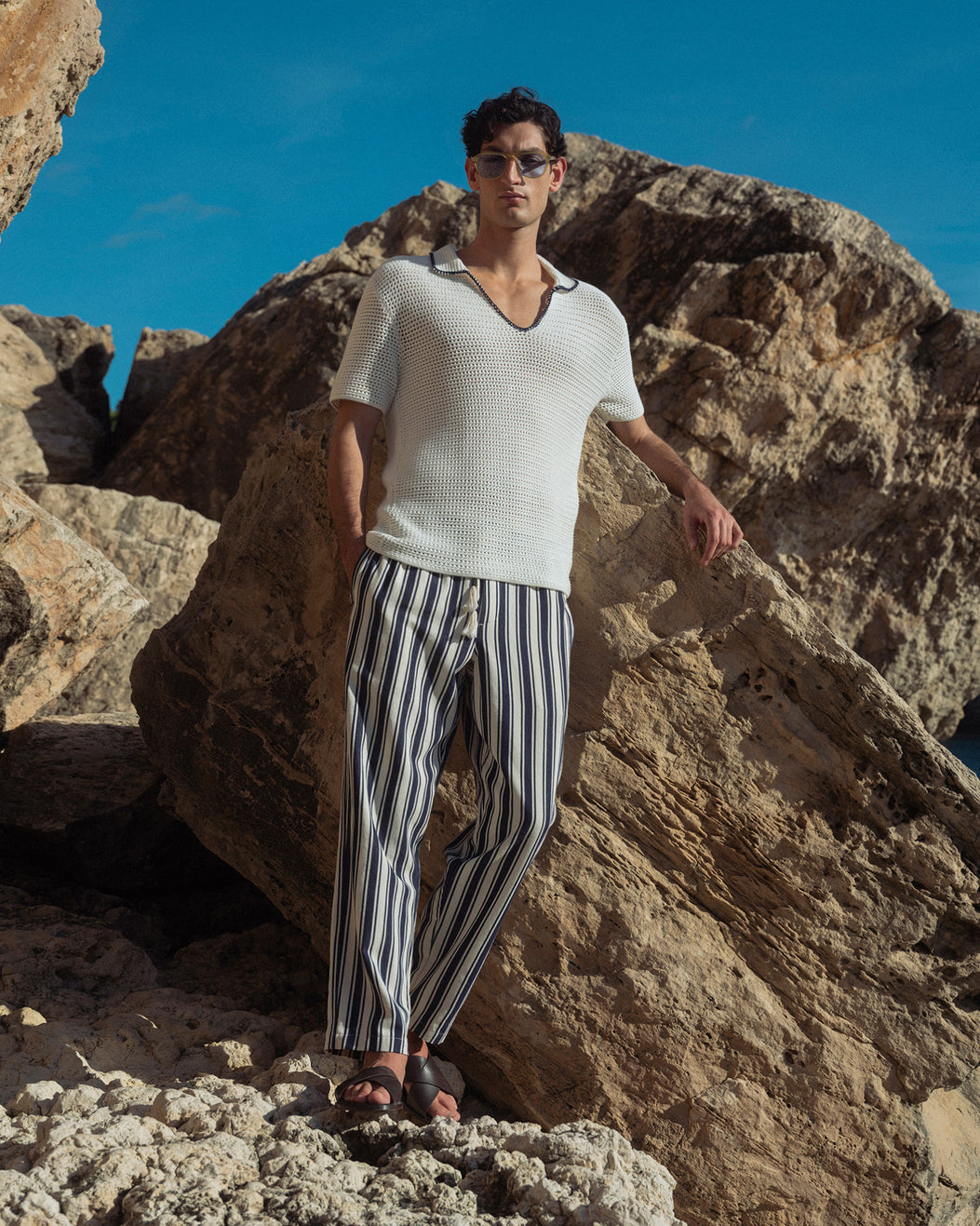 Man standing on rocky terrain wearing a white sweater and striped pants with a clear blue sky.
