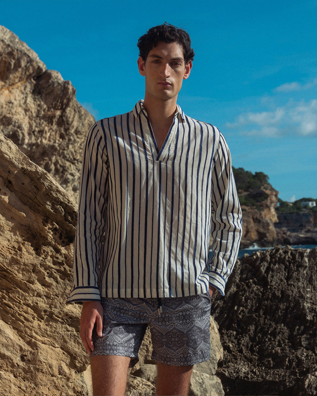 Man wearing a striped shirt and shorts standing on rocky terrain with a blue sky.