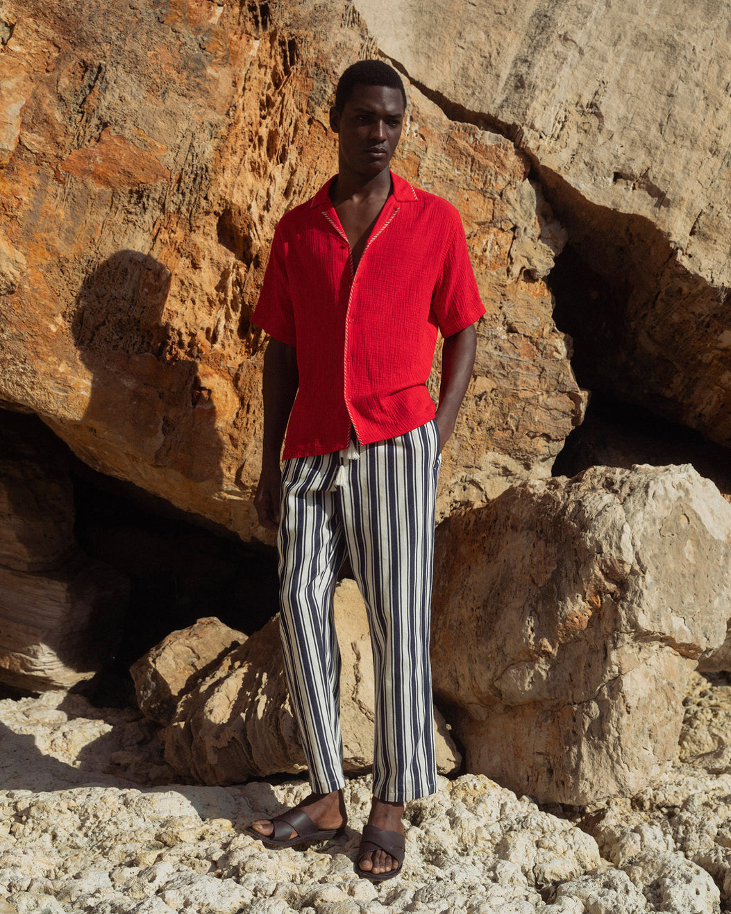 Man wearing a red shirt and striped pants standing against a rocky background