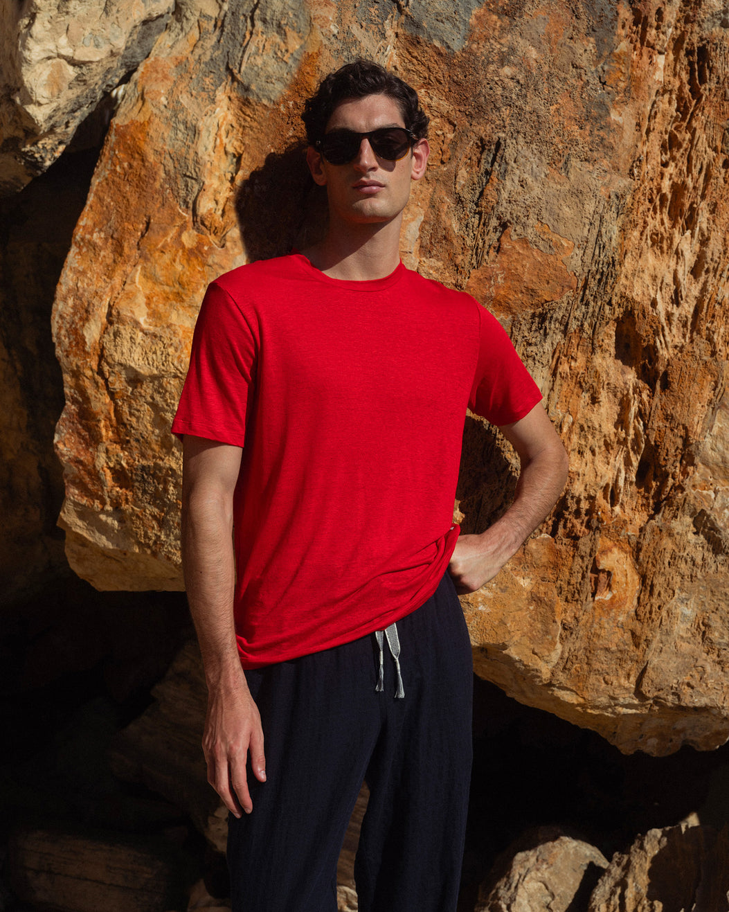 Man wearing a red shirt and sunglasses standing against a rocky background