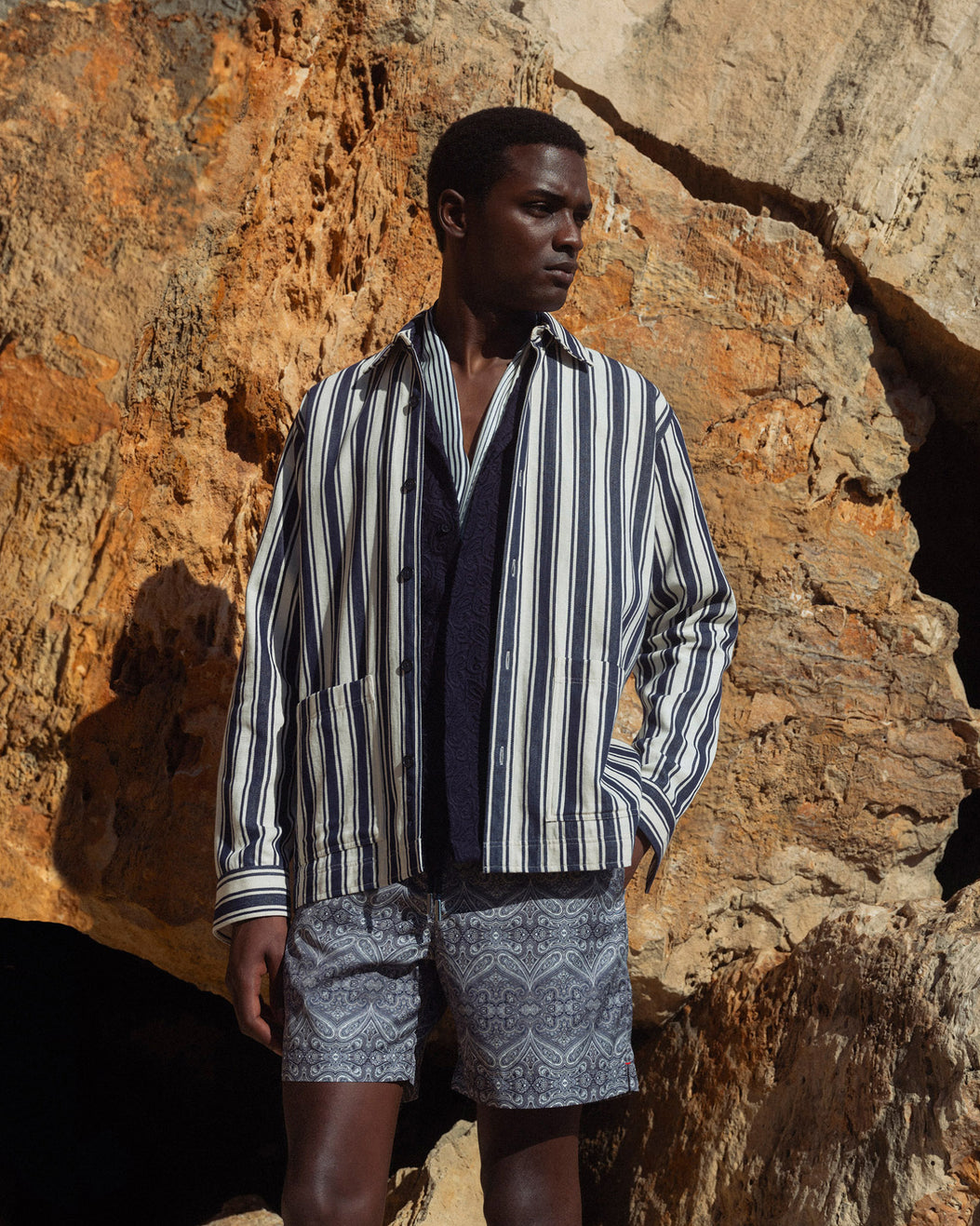 Man wearing a striped shirt and patterned shorts standing against a rocky wall.