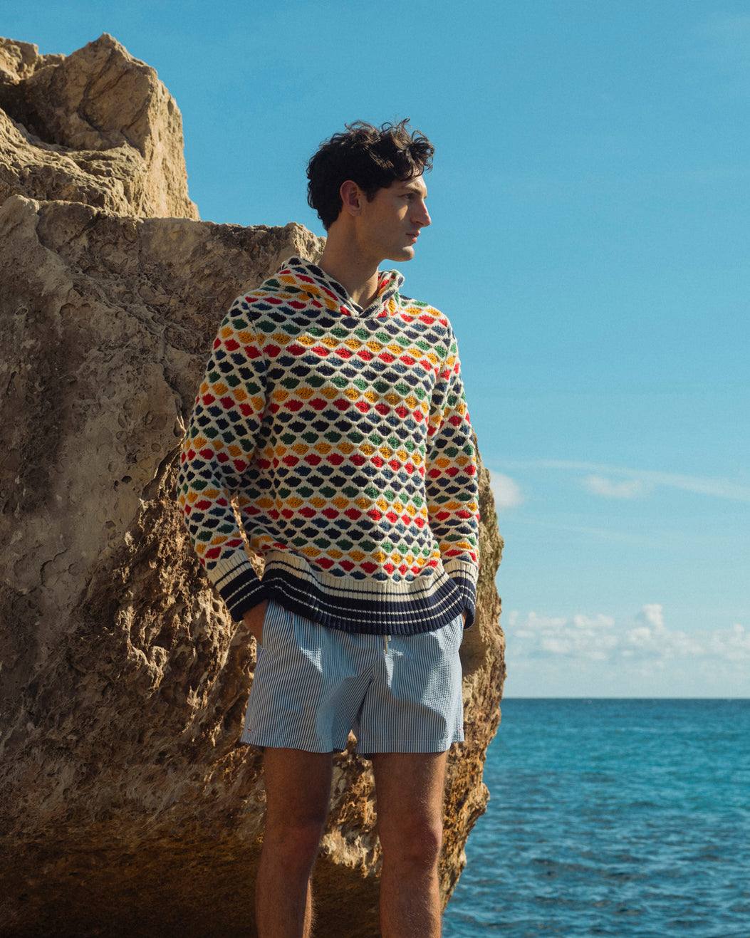 Man wearing a colorful patterned sweater and light shorts standing by a rocky coastline with blue sky and ocean.
