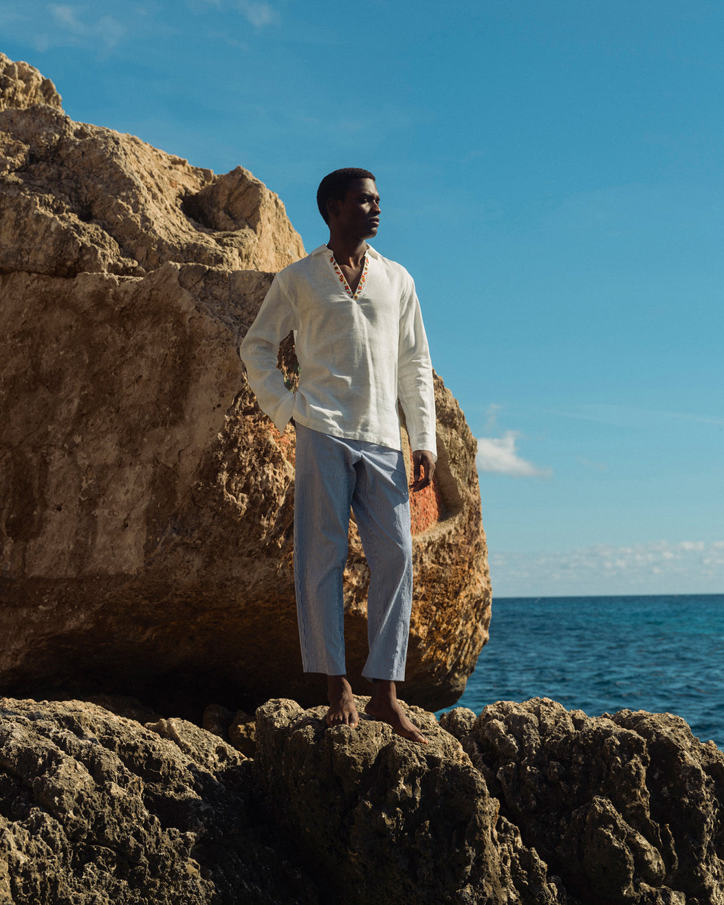 Man in a white shirt and light pants standing on rocks by the ocean with a clear blue sky.
