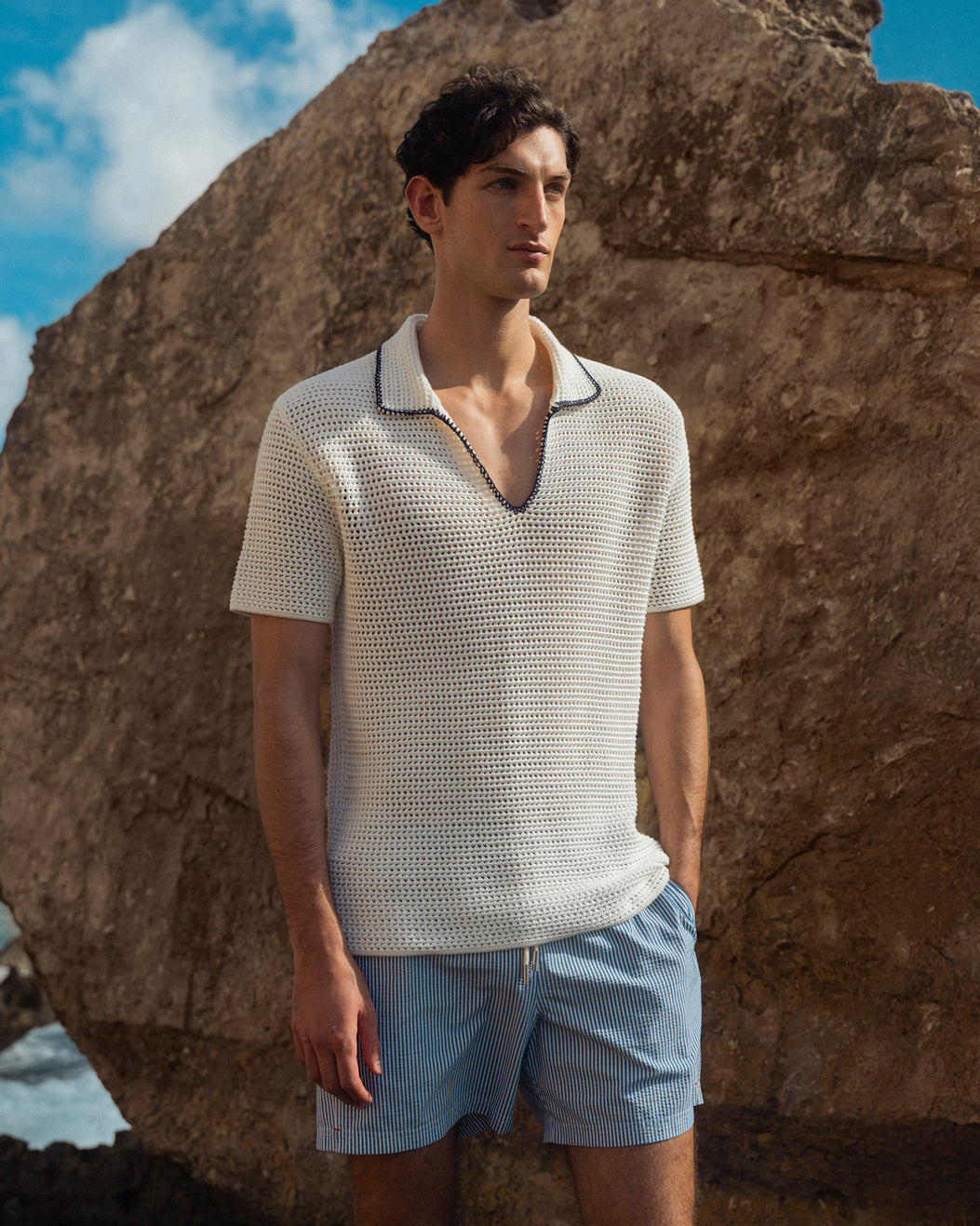 Man wearing a textured white shirt and light blue shorts standing against a rocky background.