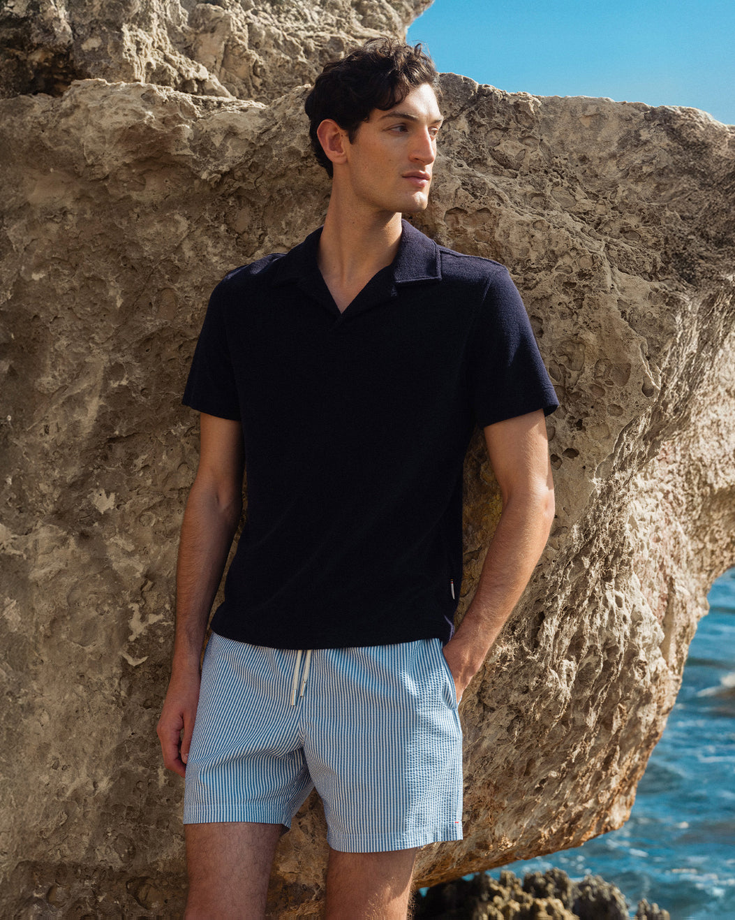 Man wearing a black polo shirt and light blue shorts standing against a rocky background with water.