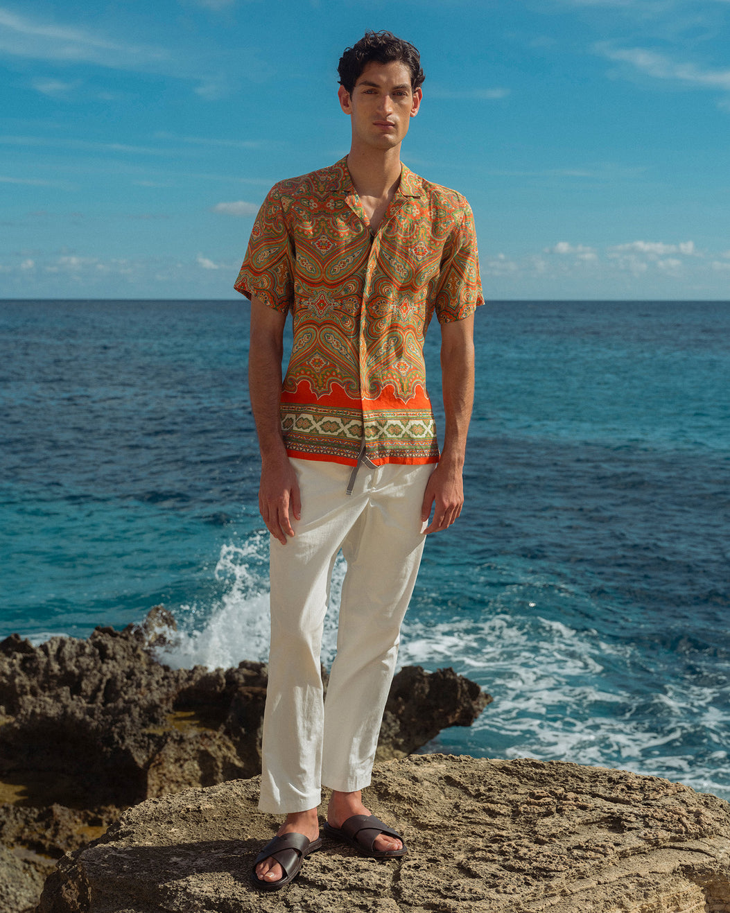 Man wearing a patterned shirt and white pants standing on a rocky coastline with ocean in the background