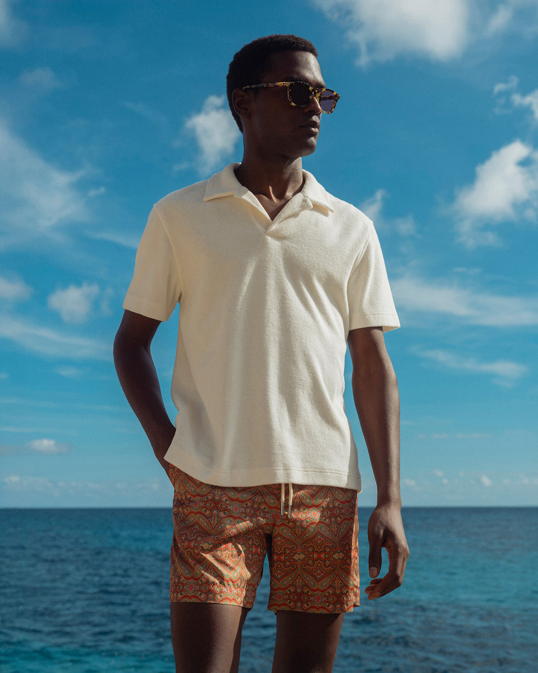 Man wearing a cream polo shirt and patterned shorts standing against a blue sky and ocean.